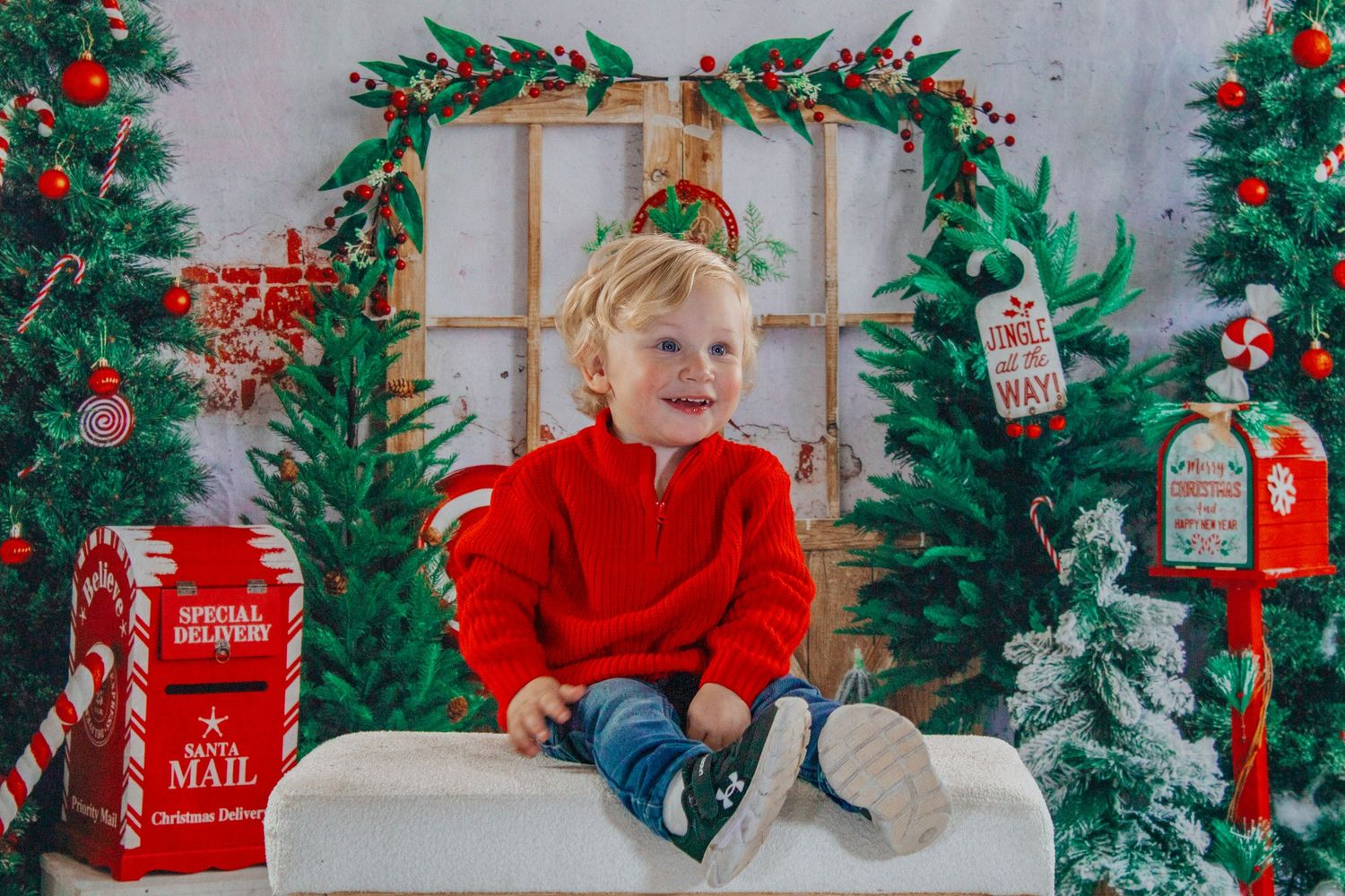 A smiling toddler in a red sweater sits on a bench surrounded by Christmas trees and holiday decorations.