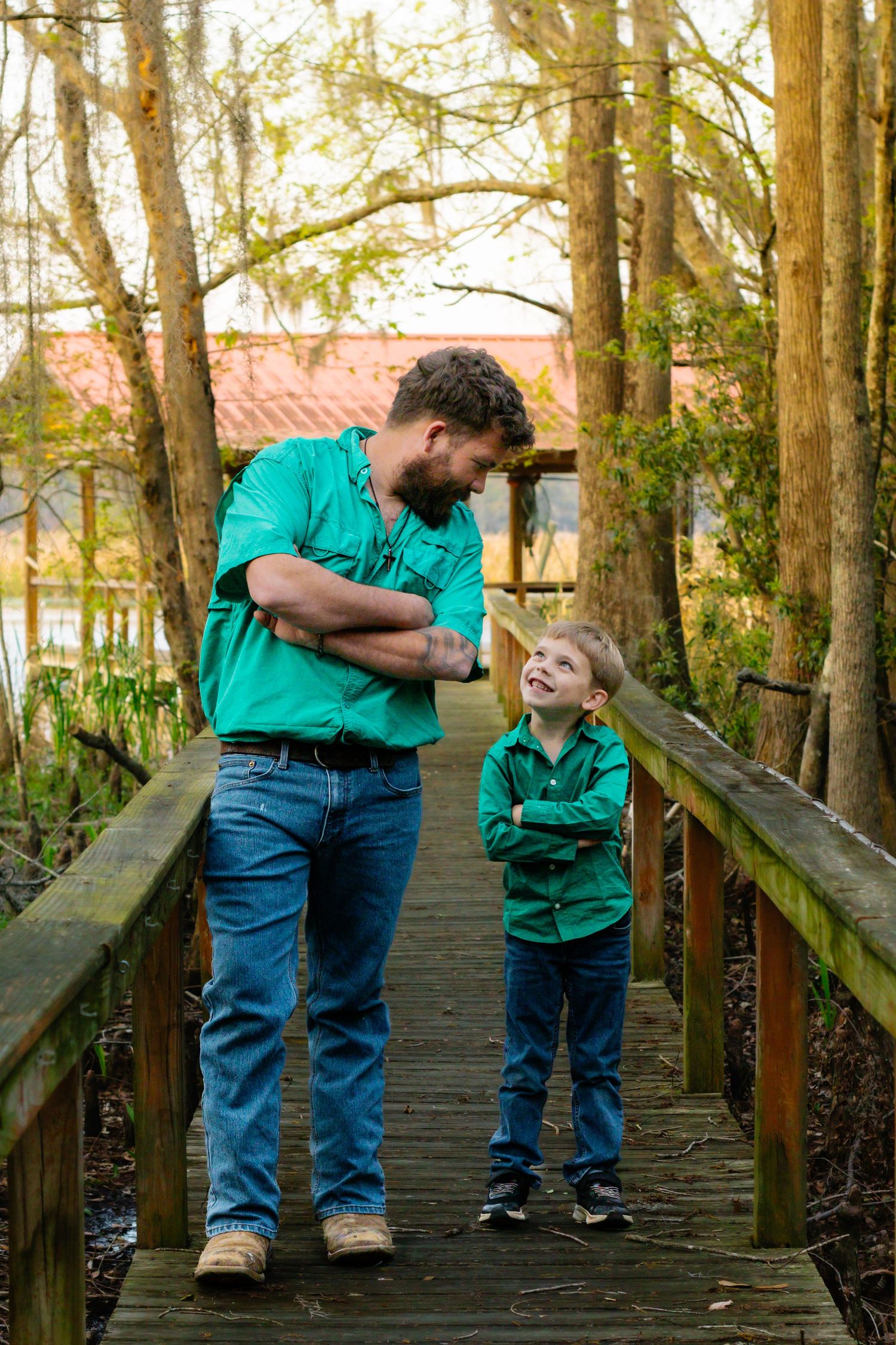 Father and son in matching green shirts stand on a wooden bridge smiling at each other.