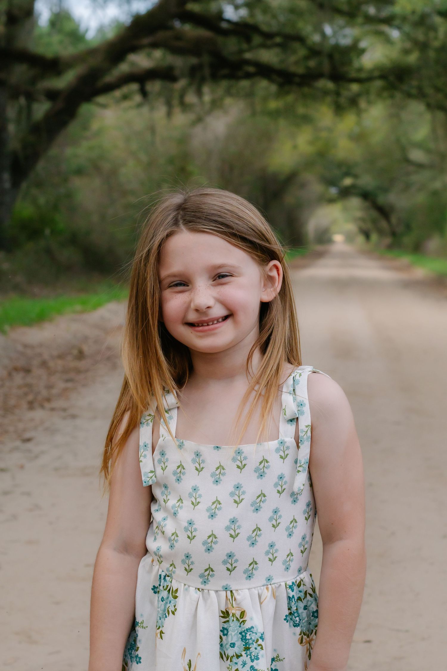 A smiling young girl in a floral sundress stands on a scenic dirt road.