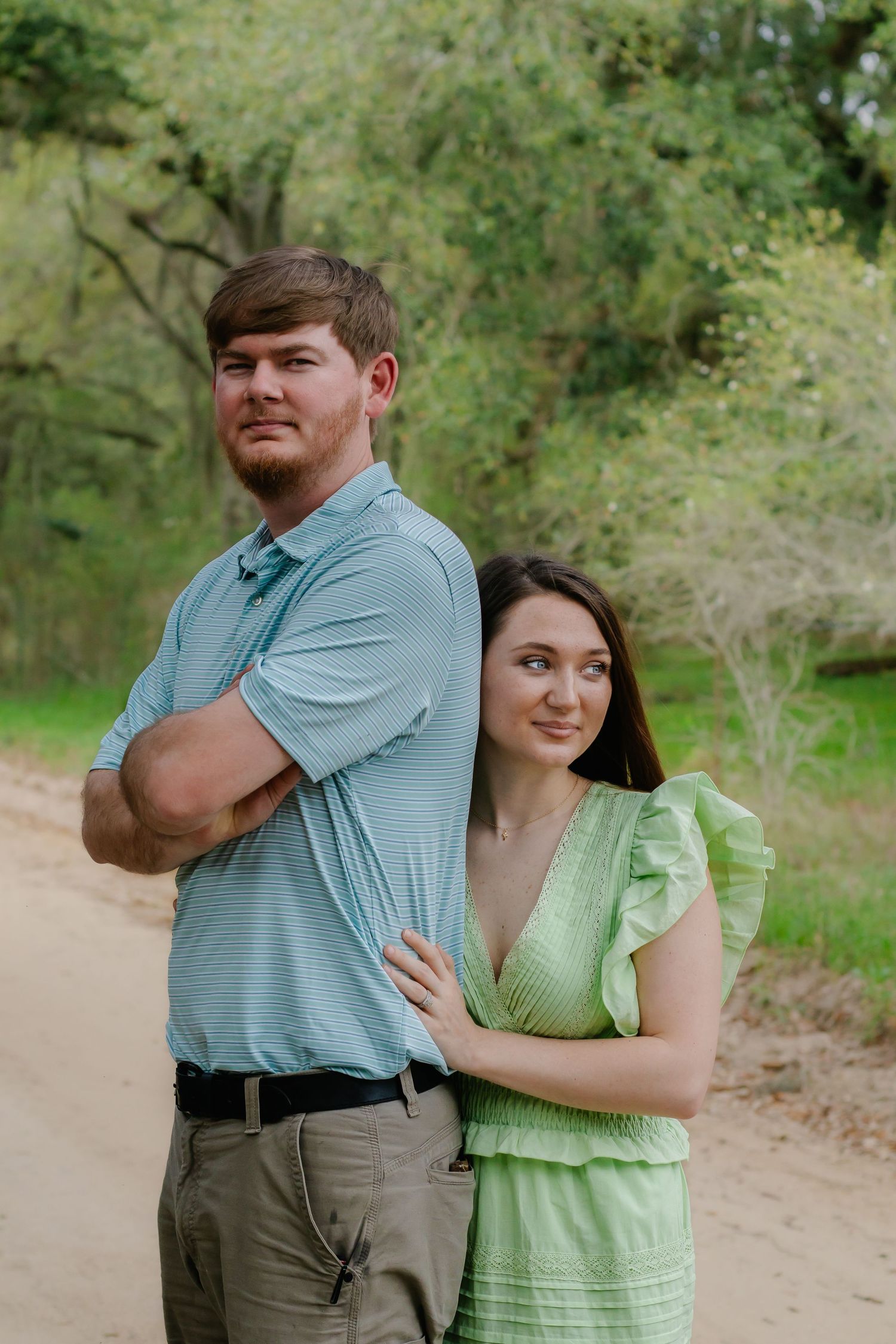 Couple posing back-to-back on a dirt road, man in blue polo with arms crossed, woman in green dress.