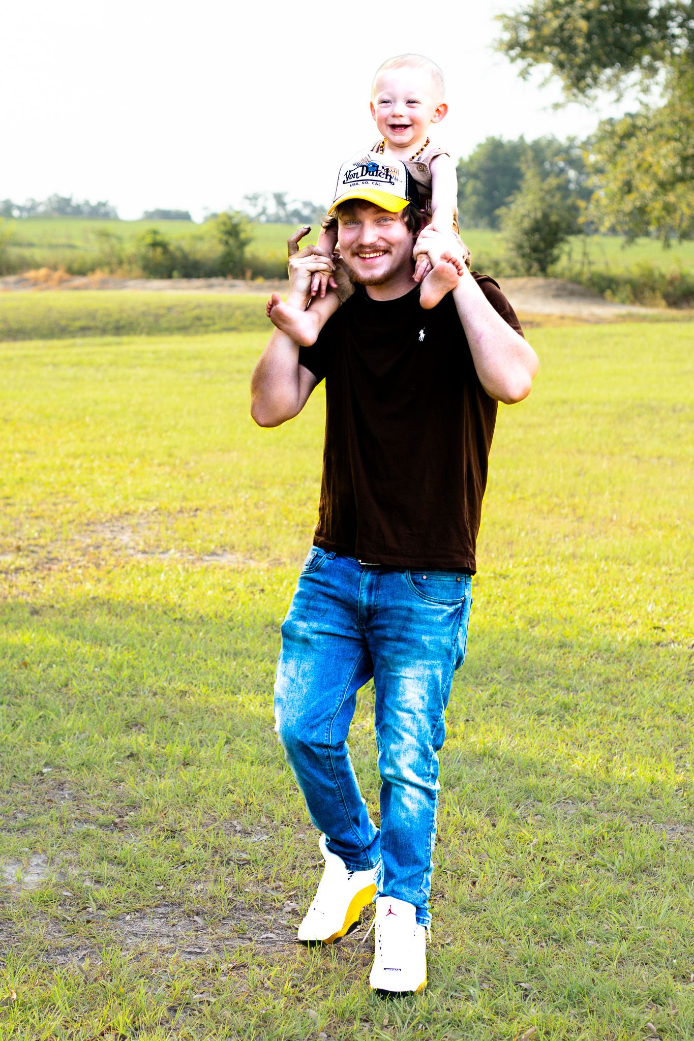 Smiling man carrying happy toddler on shoulders walking in sunny green field.