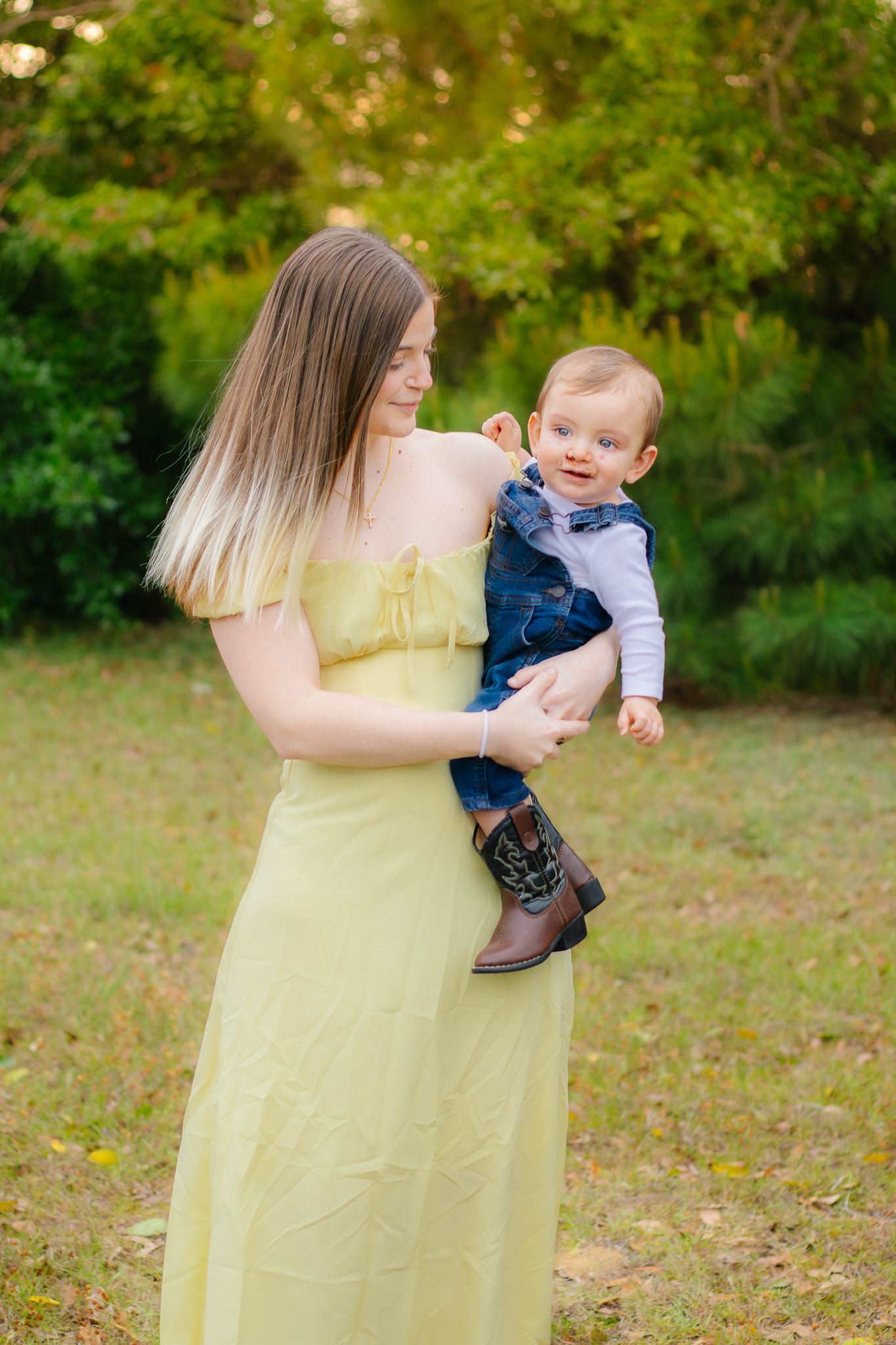 A young woman in a yellow dress holds a smiling baby in denim overalls and cowboy boots outdoors.
