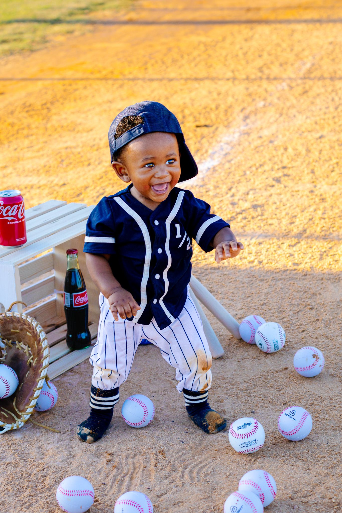 Smiling baby boy in a baseball uniform surrounded by baseballs on a dirt field with a Coca-Cola bottle nearby.
