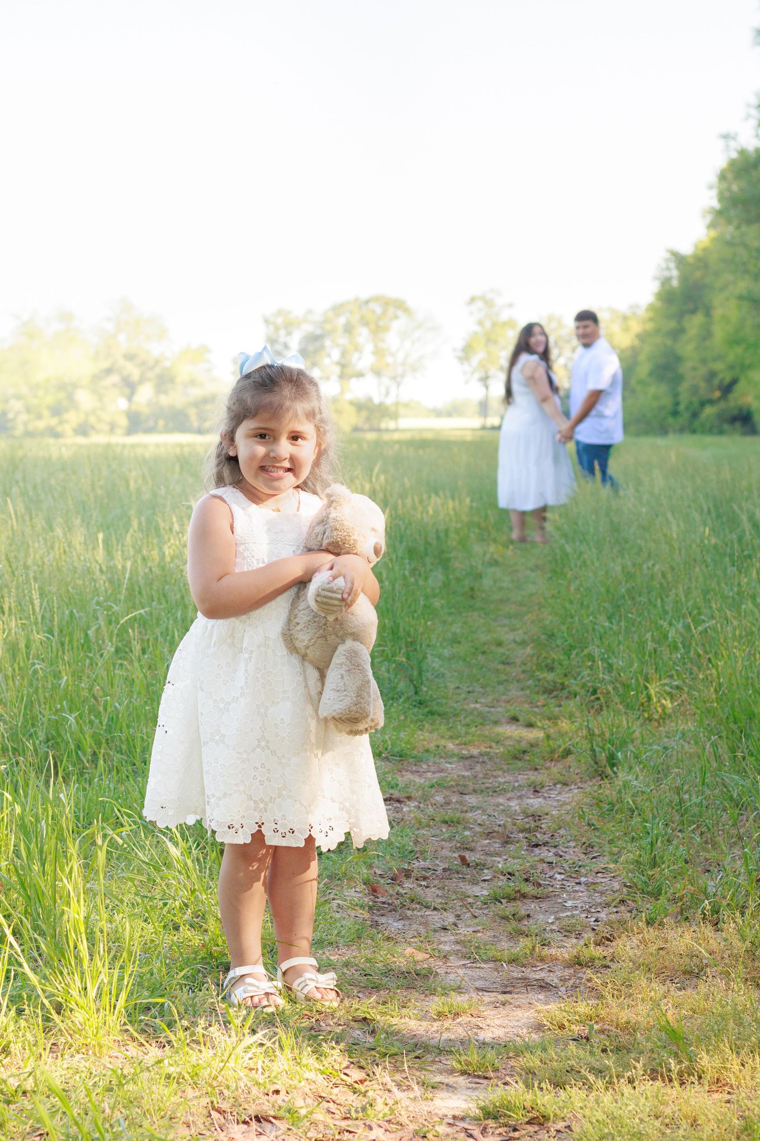 Young girl in white lace dress holds teddy bear on garden path with parents standing in background.