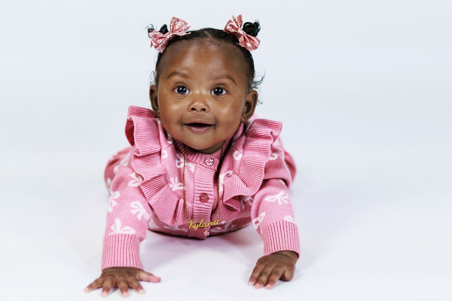 Adorable baby girl in pink ruffled outfit with bow pigtails crawling on white background.