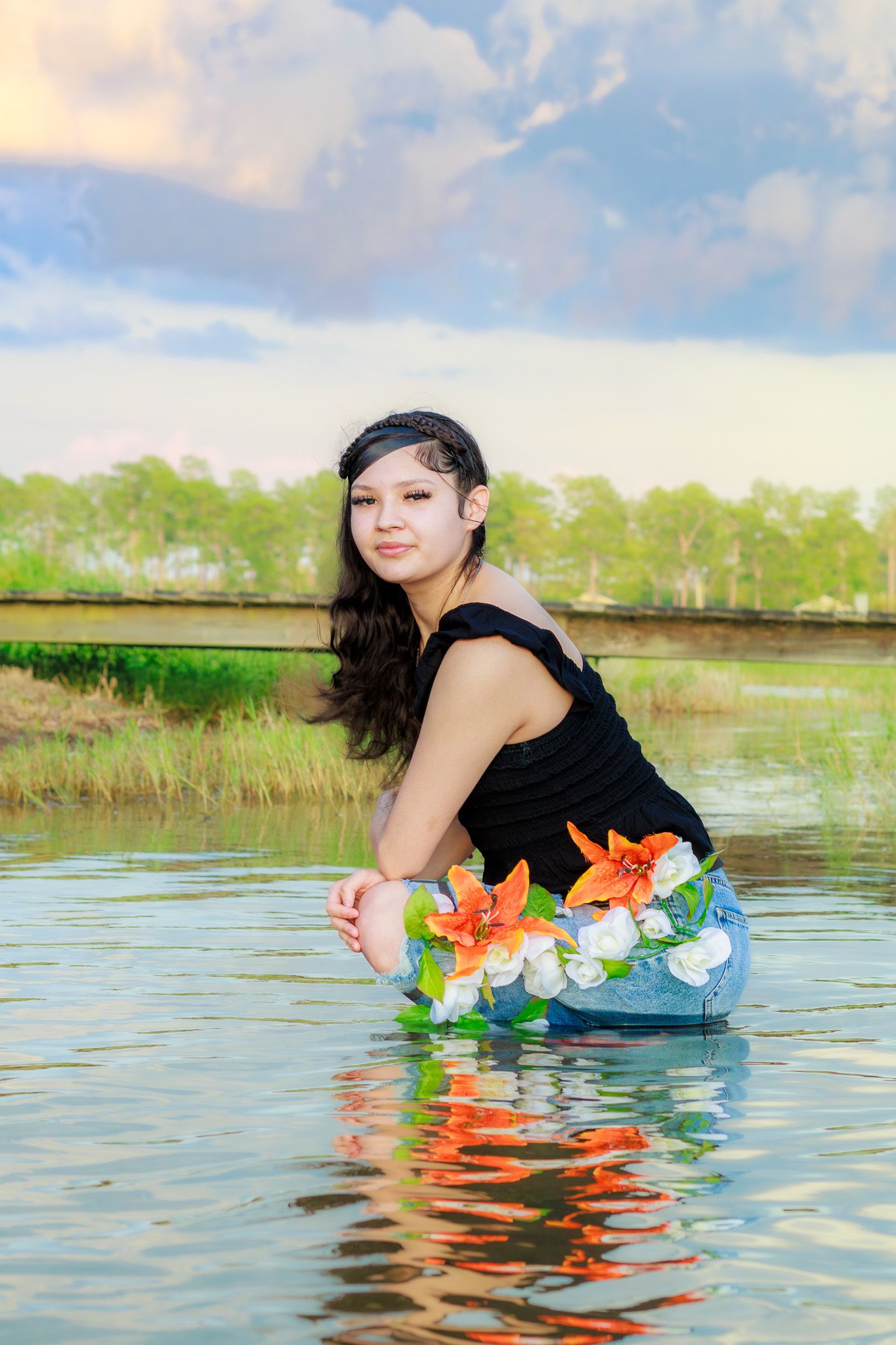 Young woman in black top and floral jeans kneeling in calm lake water at sunset.