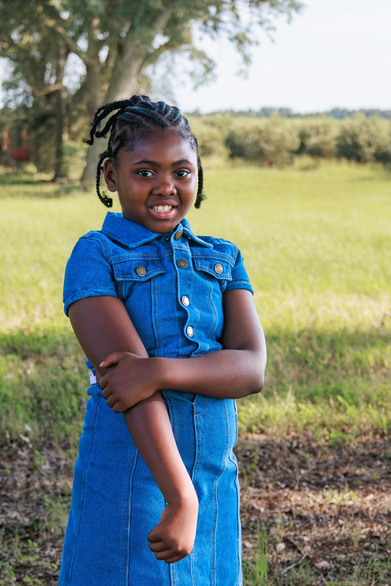 A cheerful young African girl in a blue denim dress stands with arms crossed in a sunny outdoor field.