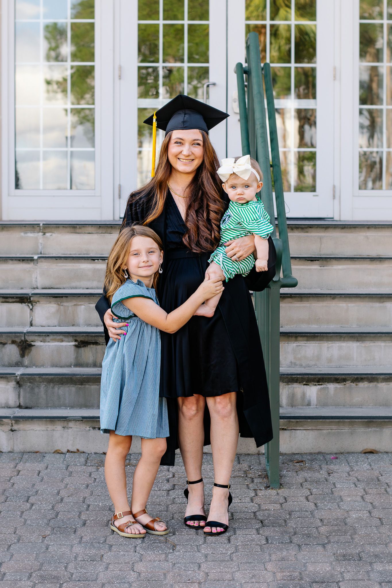 A proud graduate in cap and gown poses with her two young daughters outside a classic building.
