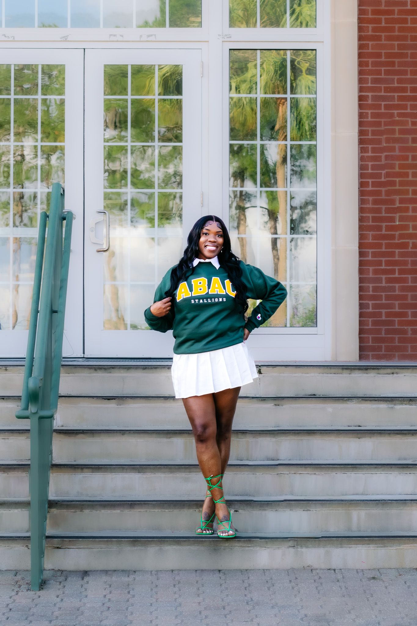 Young woman smiling in green ABAC Stallions sweatshirt and white skirt, posing on steps outside brick building.