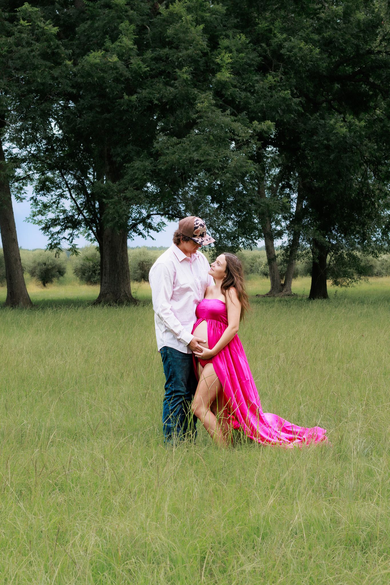 Couple embracing in a lush green field, woman in flowing pink maternity gown, romantic outdoor maternity photo session.