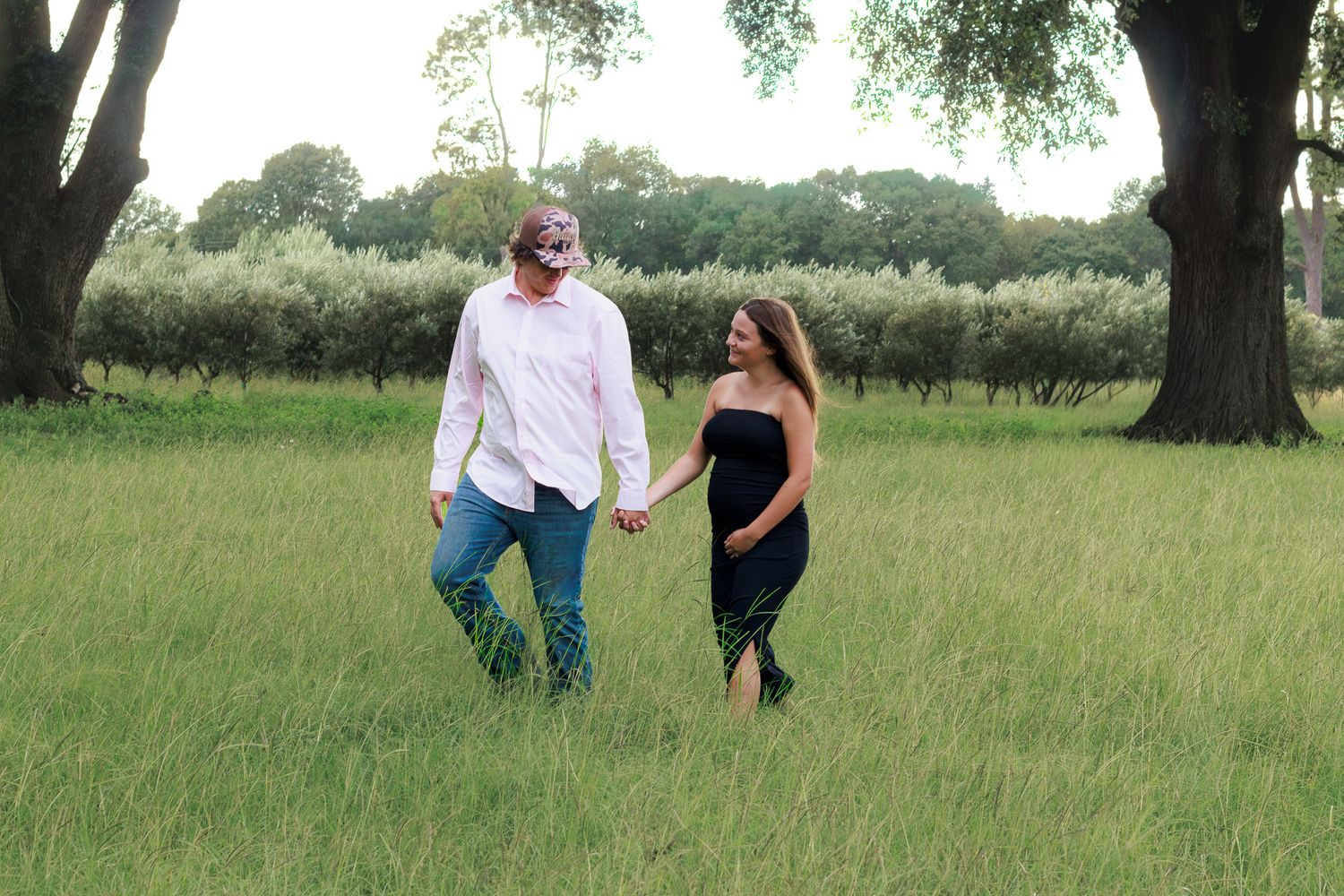 Couple holding hands walking through lush green field surrounded by trees during outdoor maternity photoshoot.