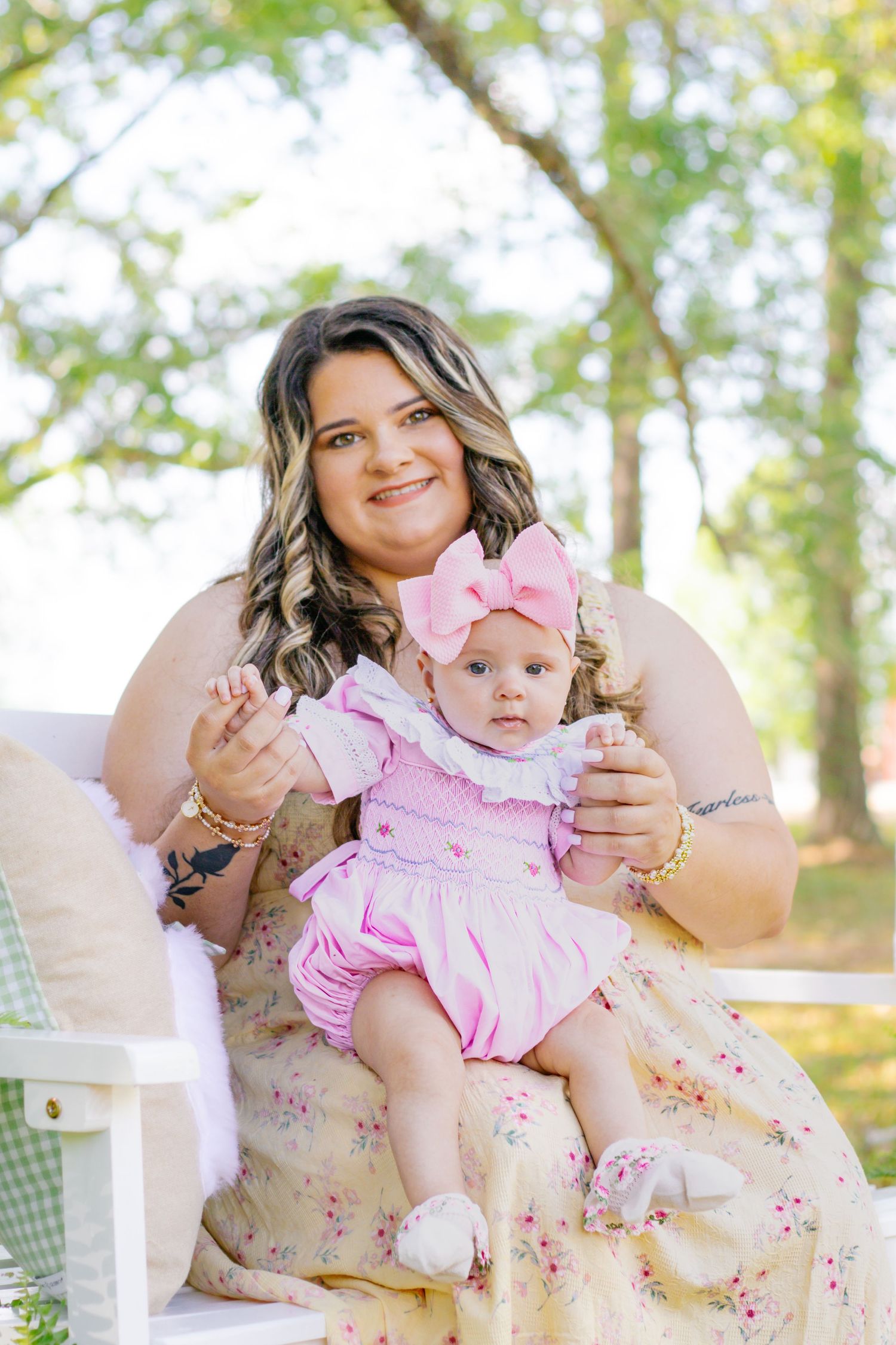 Happy mother in yellow floral dress holds adorable baby in pink bow headband outdoors.