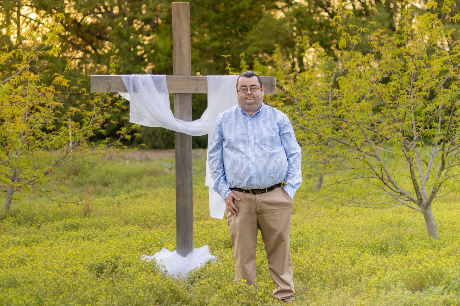 Man in blue shirt and khakis standing beside a wooden cross with white cloth in a green field.