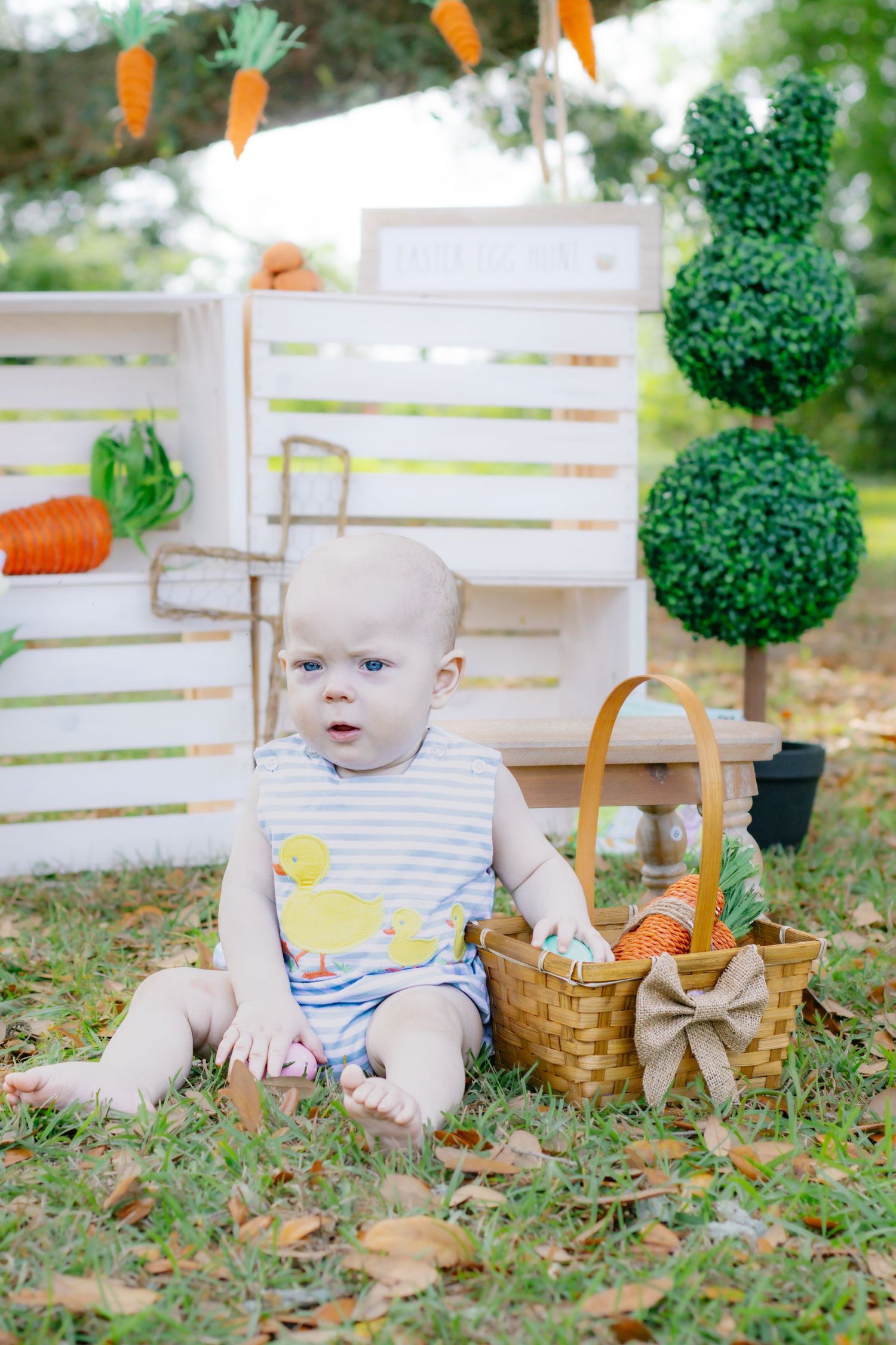 Baby boy in duck romper sits on grass beside Easter basket and bunny topiary decorations.