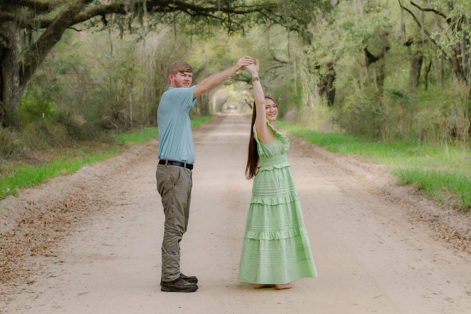 Young couple twirling together on a picturesque canopy-lined dirt road surrounded by lush green trees.