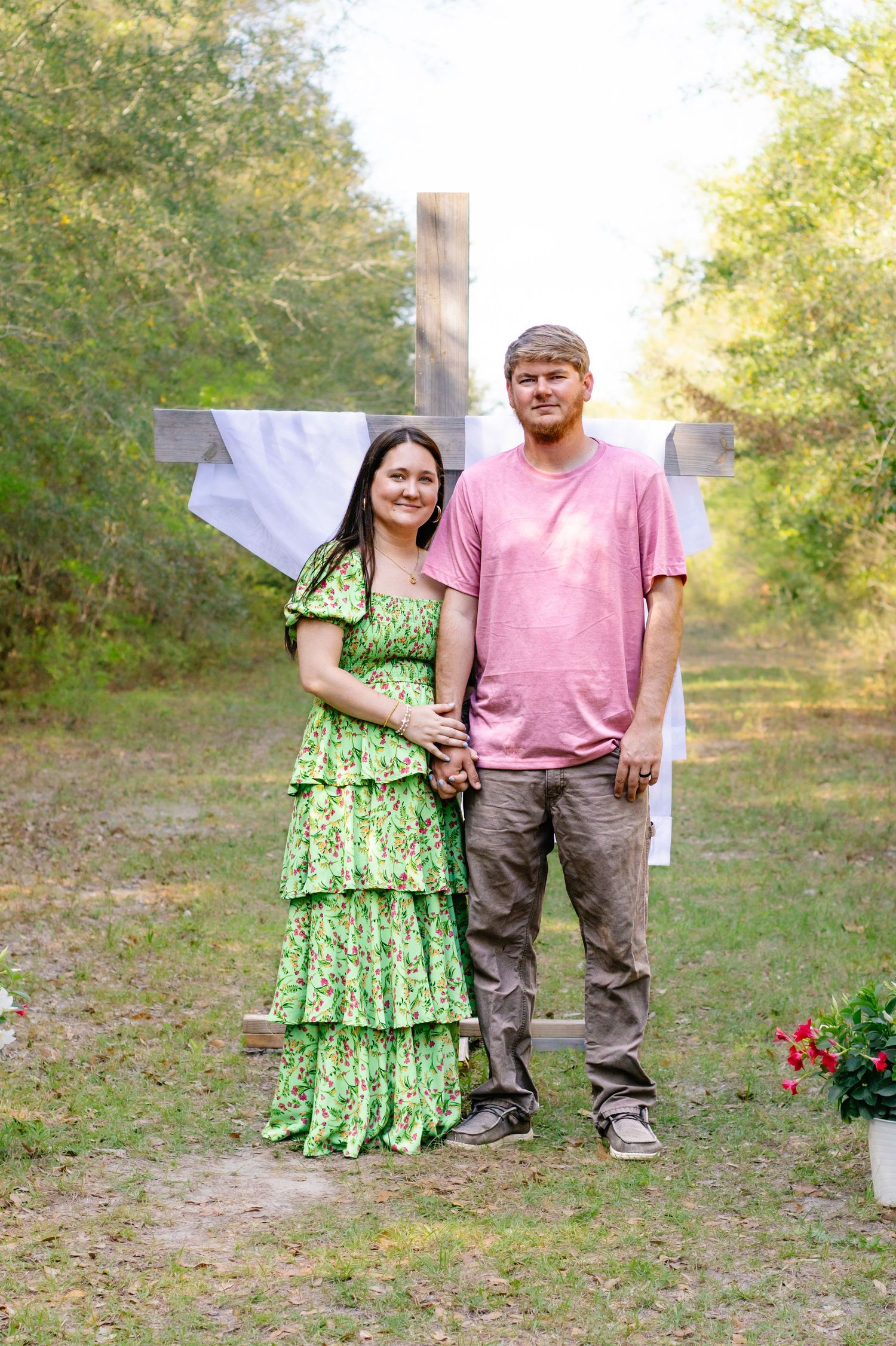 Couple holding hands in front of a large wooden cross outdoors, woman in green floral dress and man in pink shirt.