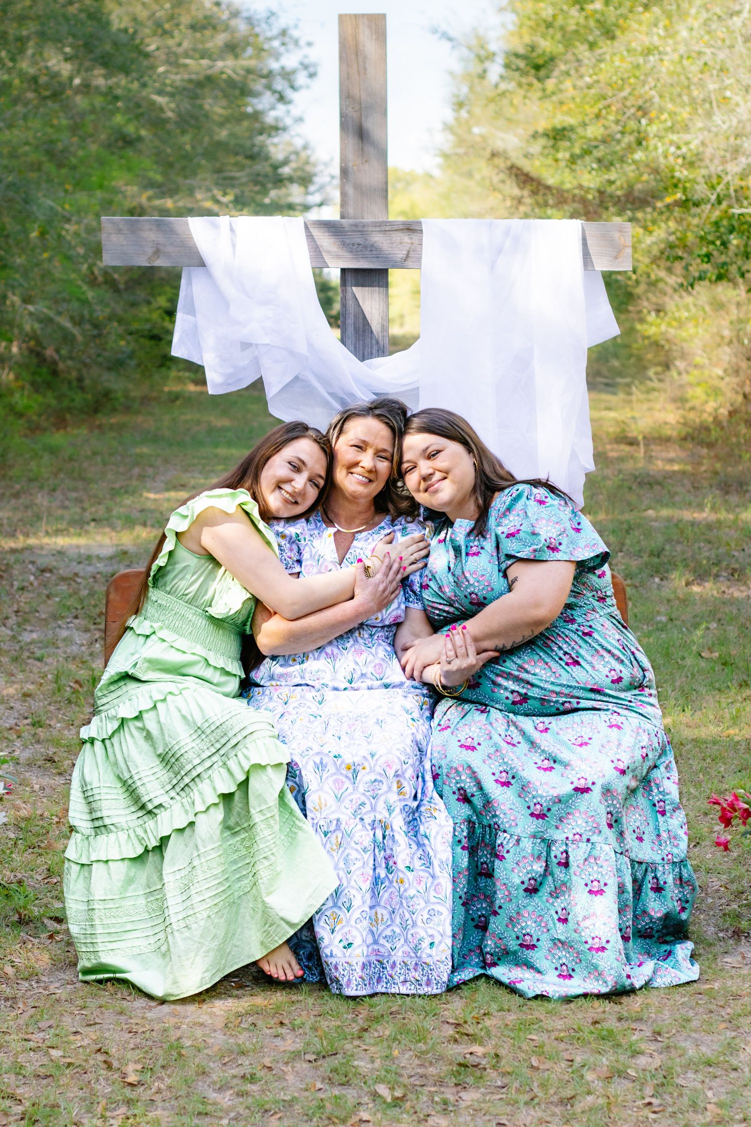 Three women in colorful floral dresses hugging and smiling together in front of a wooden cross outdoors.