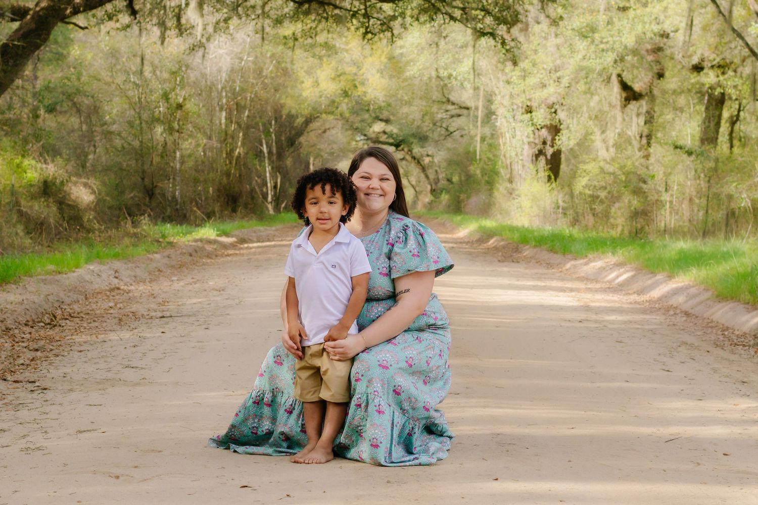 A mother in a floral dress poses with her young son on a dirt path.