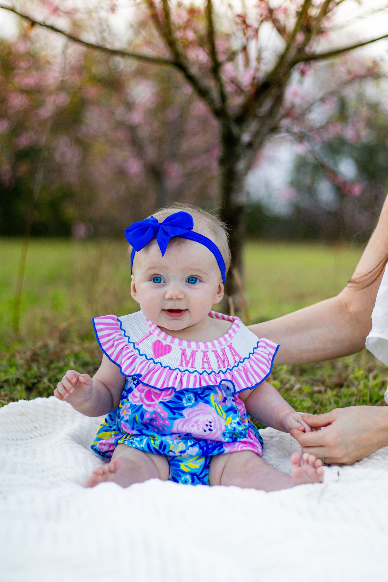 Blue-eyed baby girl in floral romper with blue bow headband sits on white blanket outdoors near pink flowering trees.