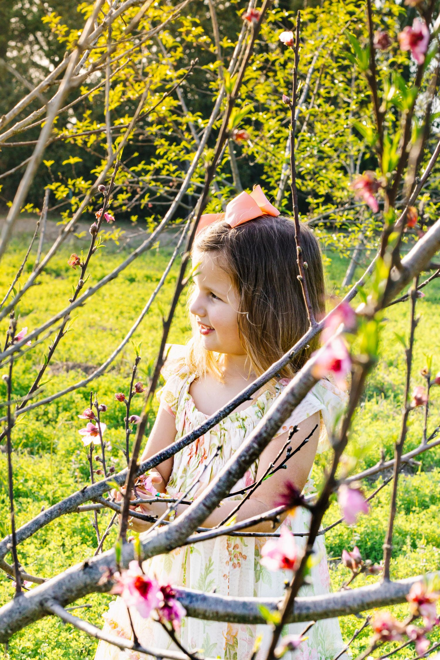 Young girl in floral dress smiling among pink blossoming tree branches in sunny spring garden.