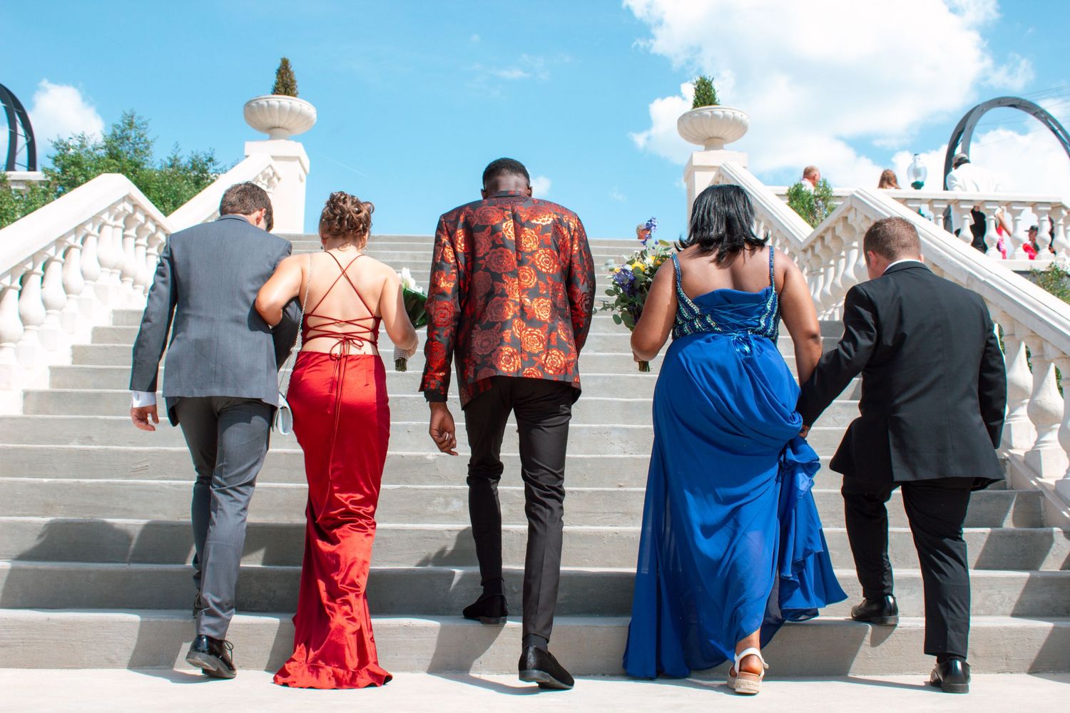 Four prom attendees walk up elegant white stairs, dressed in formal red and blue gowns and suits.