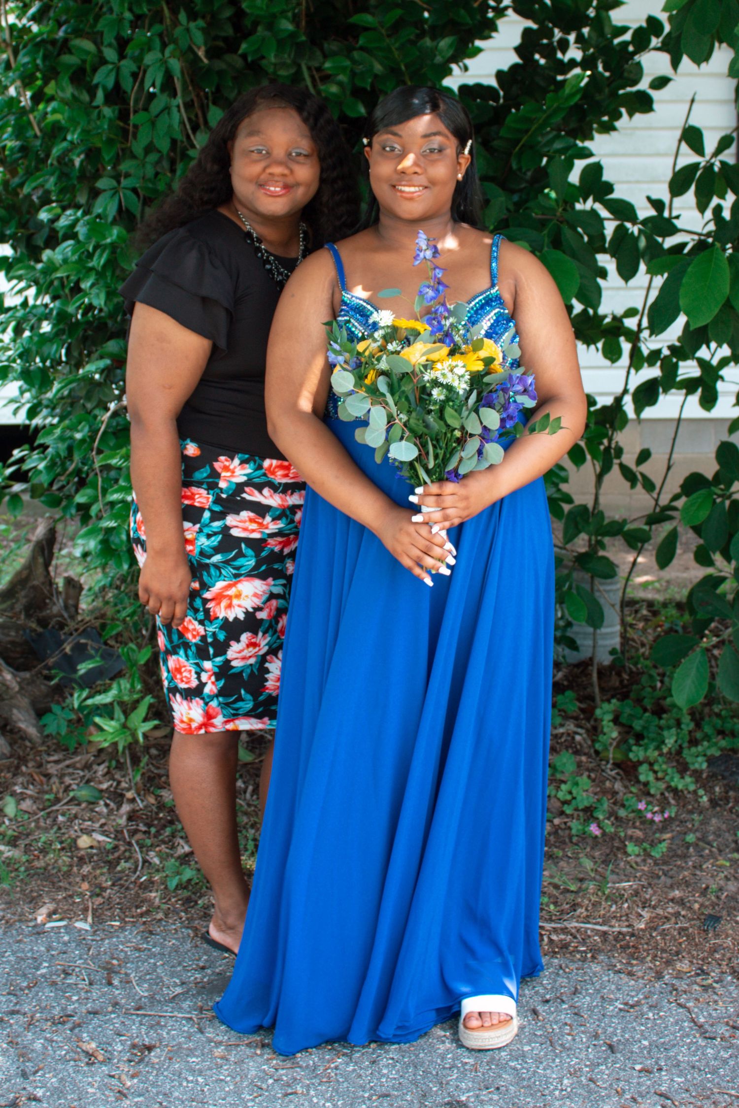 Two women pose outdoors near greenery, one in blue formal gown holding a colorful bouquet.