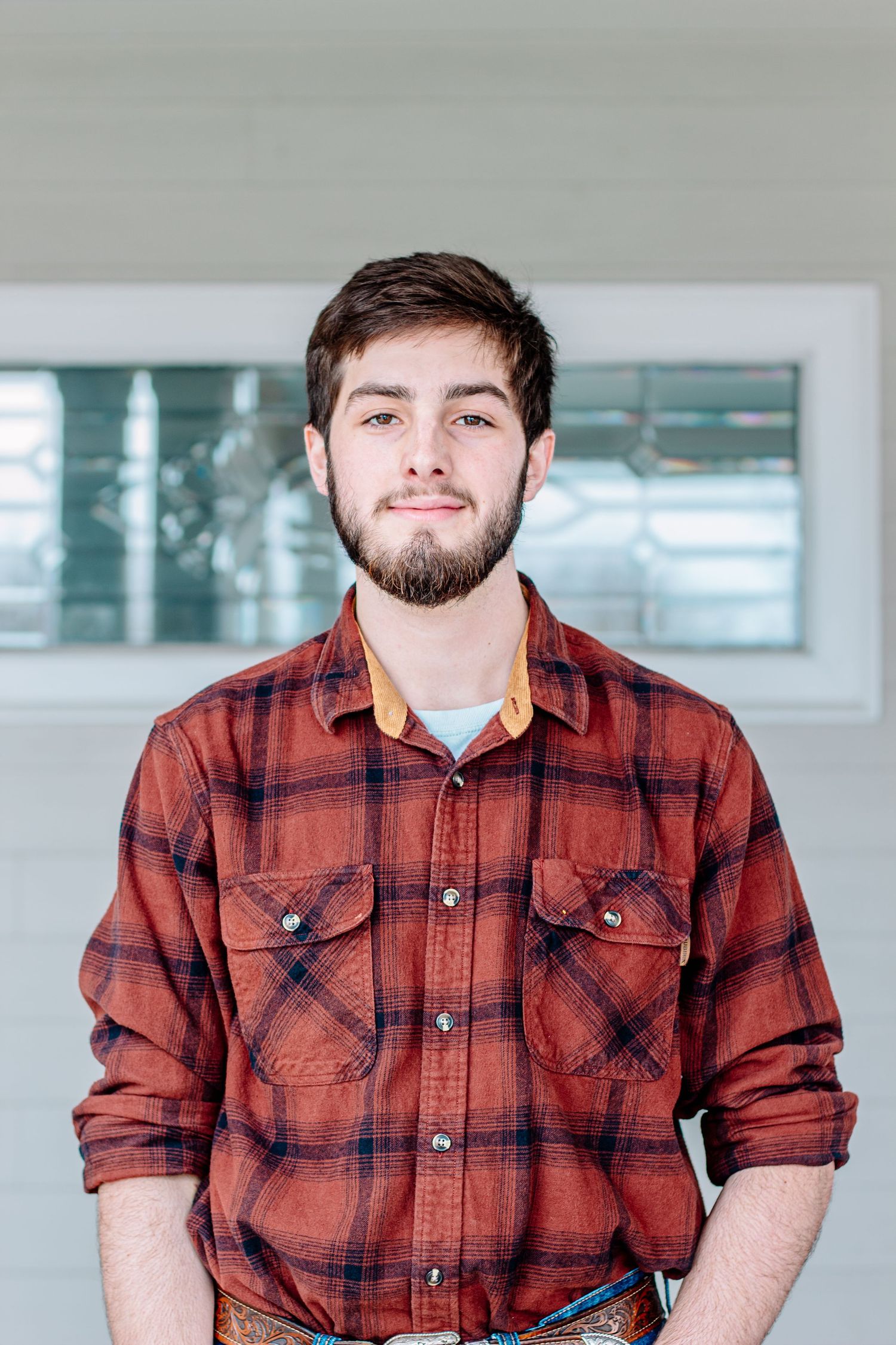 Young bearded man in a red plaid flannel shirt smiling confidently indoors.