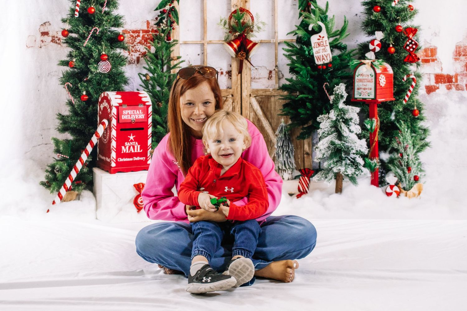 A mother in a pink hoodie sits cross-legged with her young child in a festive Christmas photo studio setting.