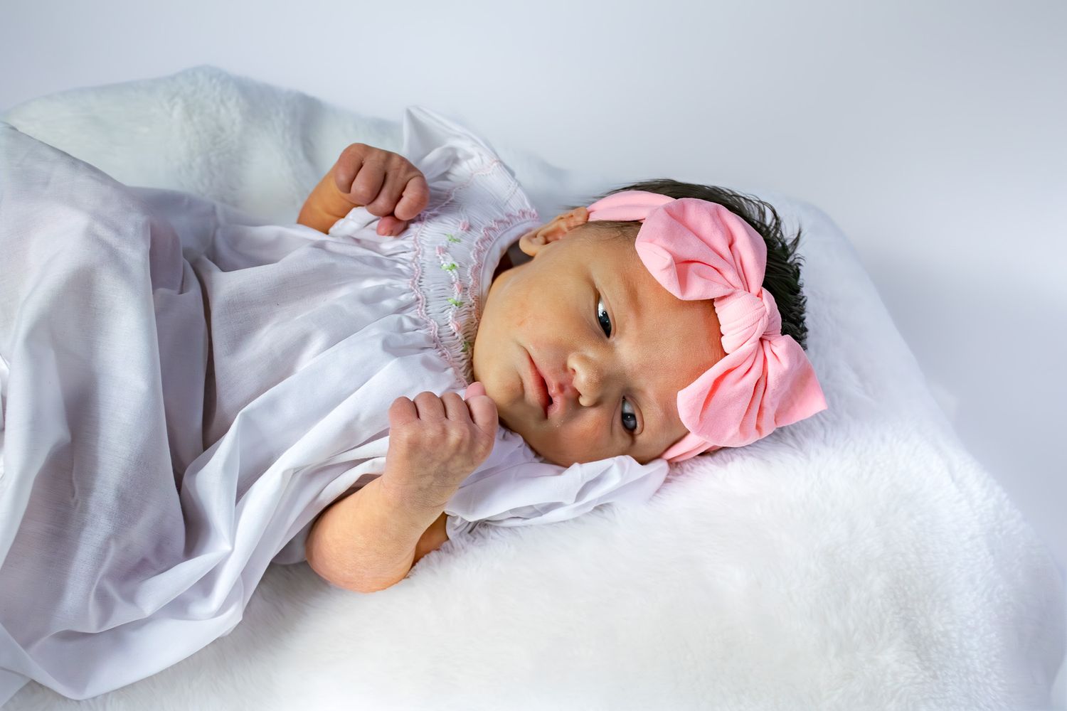 Newborn baby girl in white dress with pink bow headband lying on white fluffy blanket.