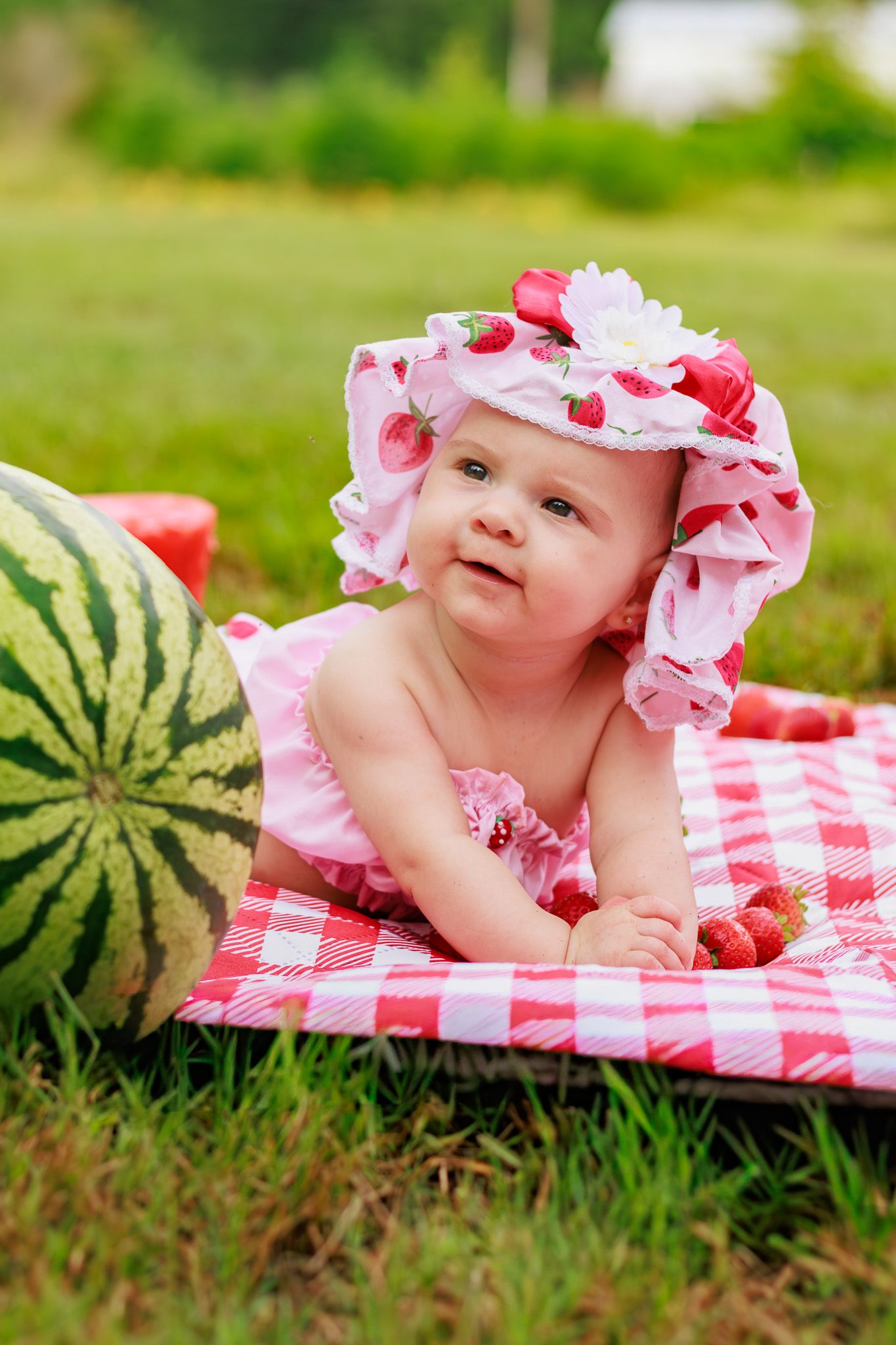 Cute baby in pink strawberry hat lies on red gingham picnic blanket next to a whole watermelon in garden.