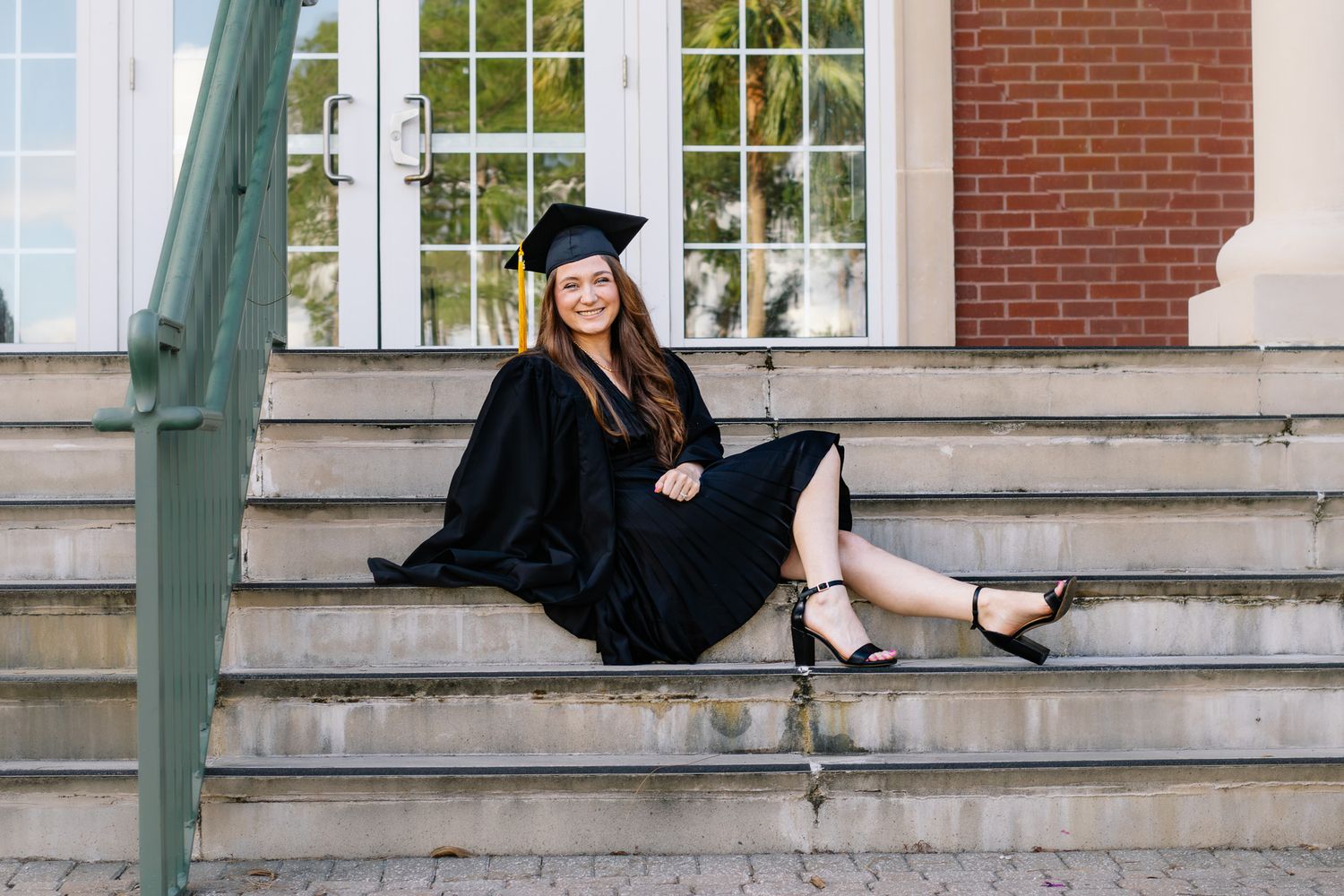 Smiling female graduate in cap and gown sits relaxed on stone steps outside brick university building.