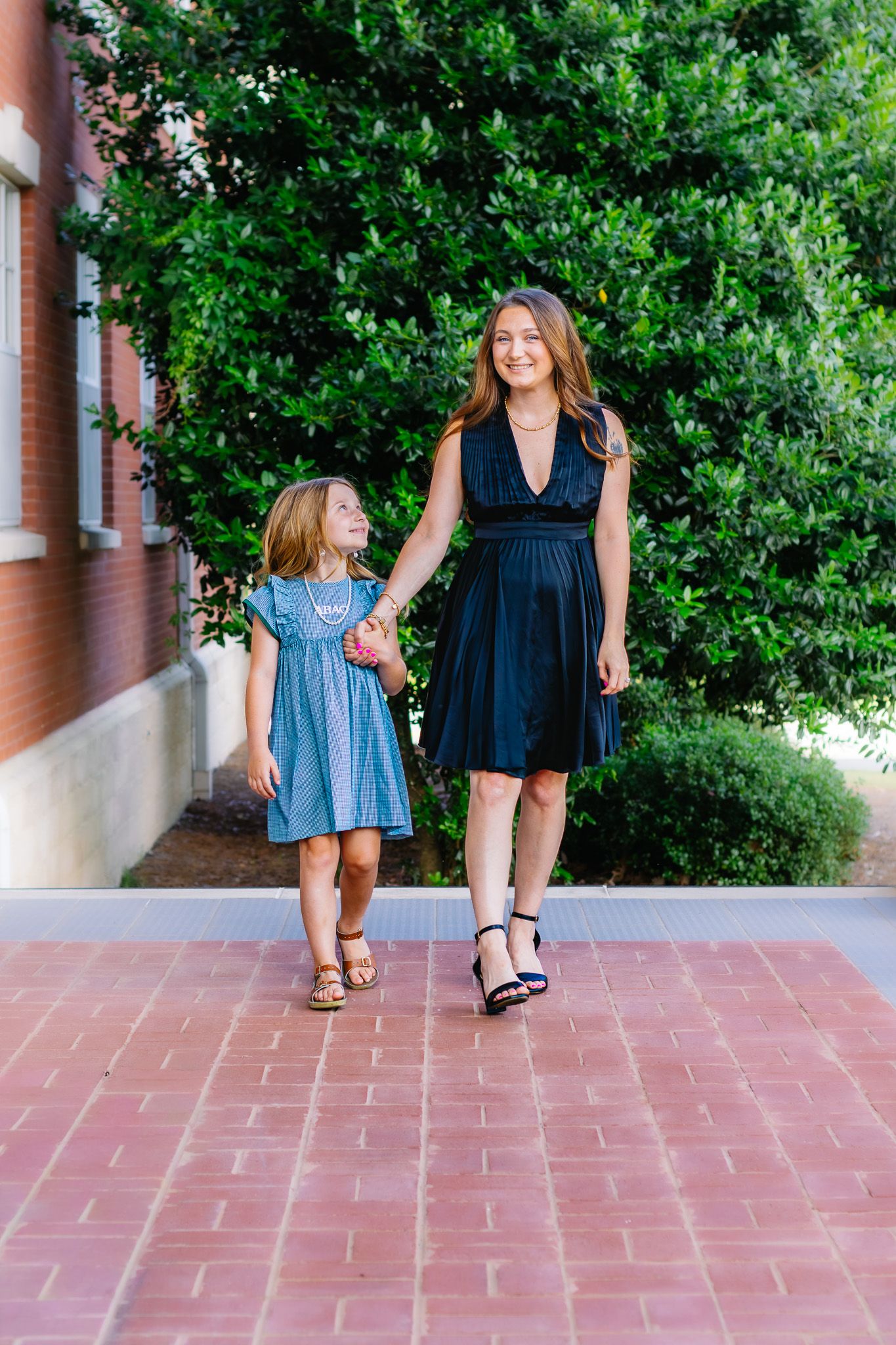 Woman in black dress and girl in blue dress walk hand-in-hand on brick path near green trees.