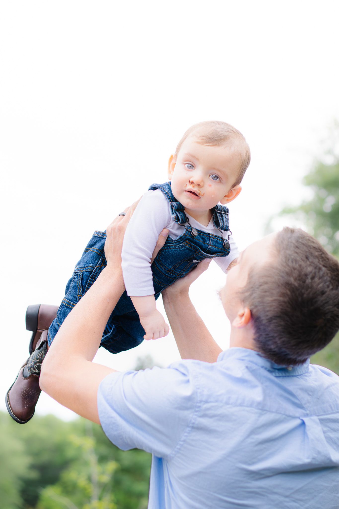Father lifting happy baby in denim overalls high in the air against bright sky outdoors.