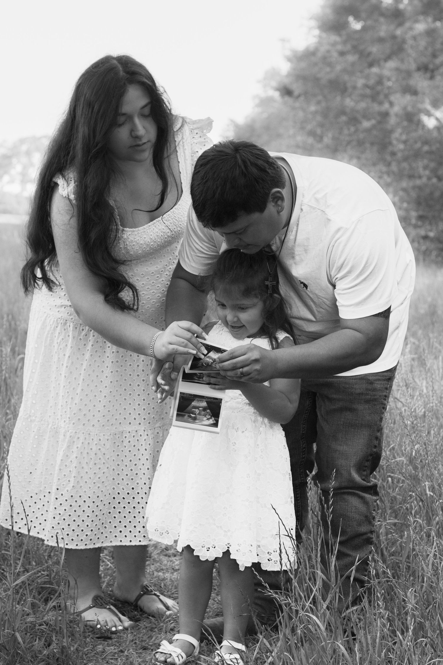 Black and white family photo of couple and toddler looking at ultrasound images in a grassy field.