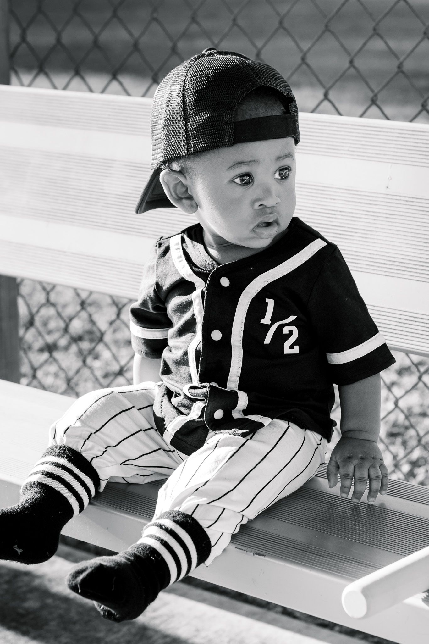 Black and white photo of a baby boy in a baseball uniform with number 1/2, sitting on a bench wearing a backwards cap.