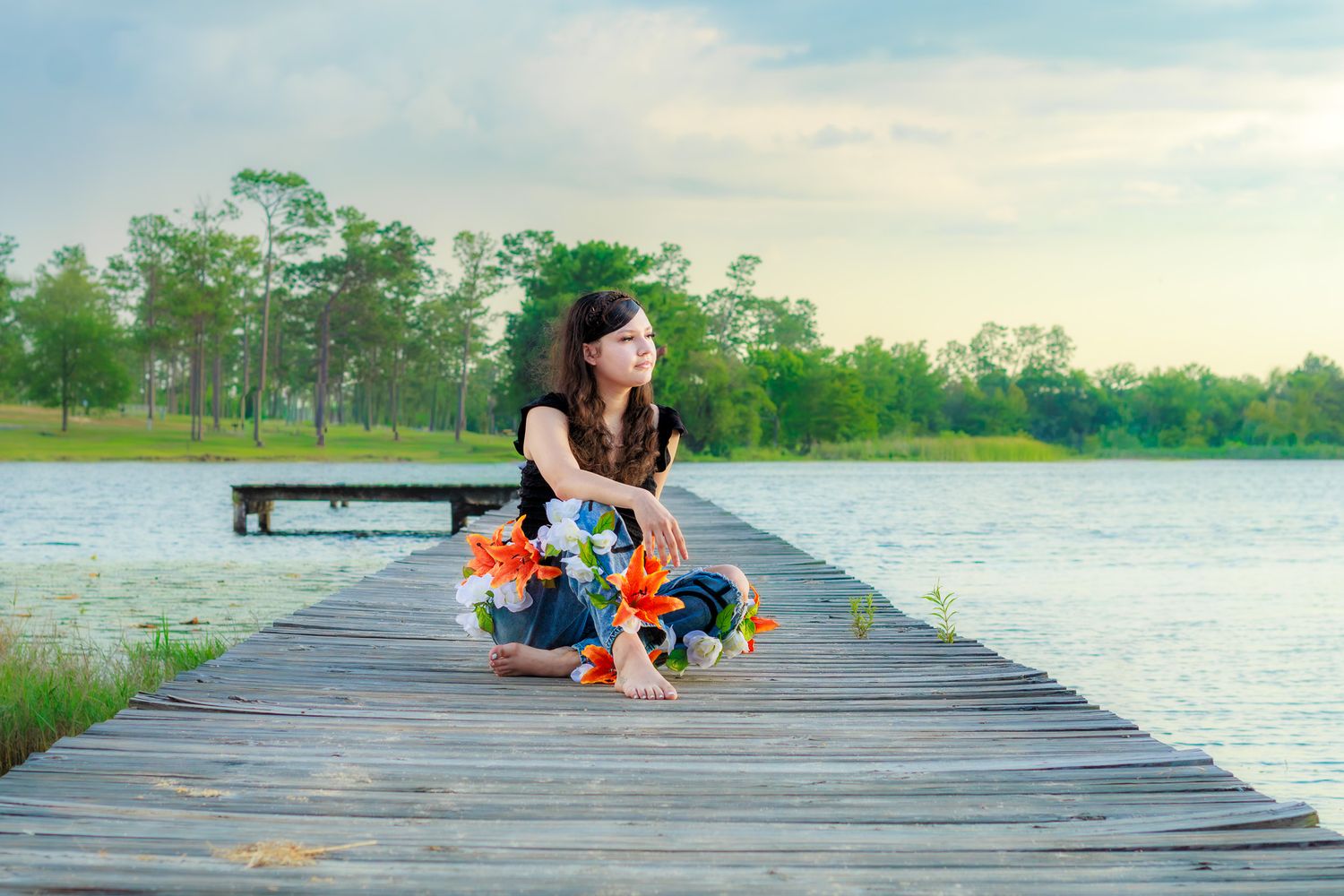 Young woman sitting on a wooden dock by a serene lake, holding colorful flowers with lush green trees in the background.