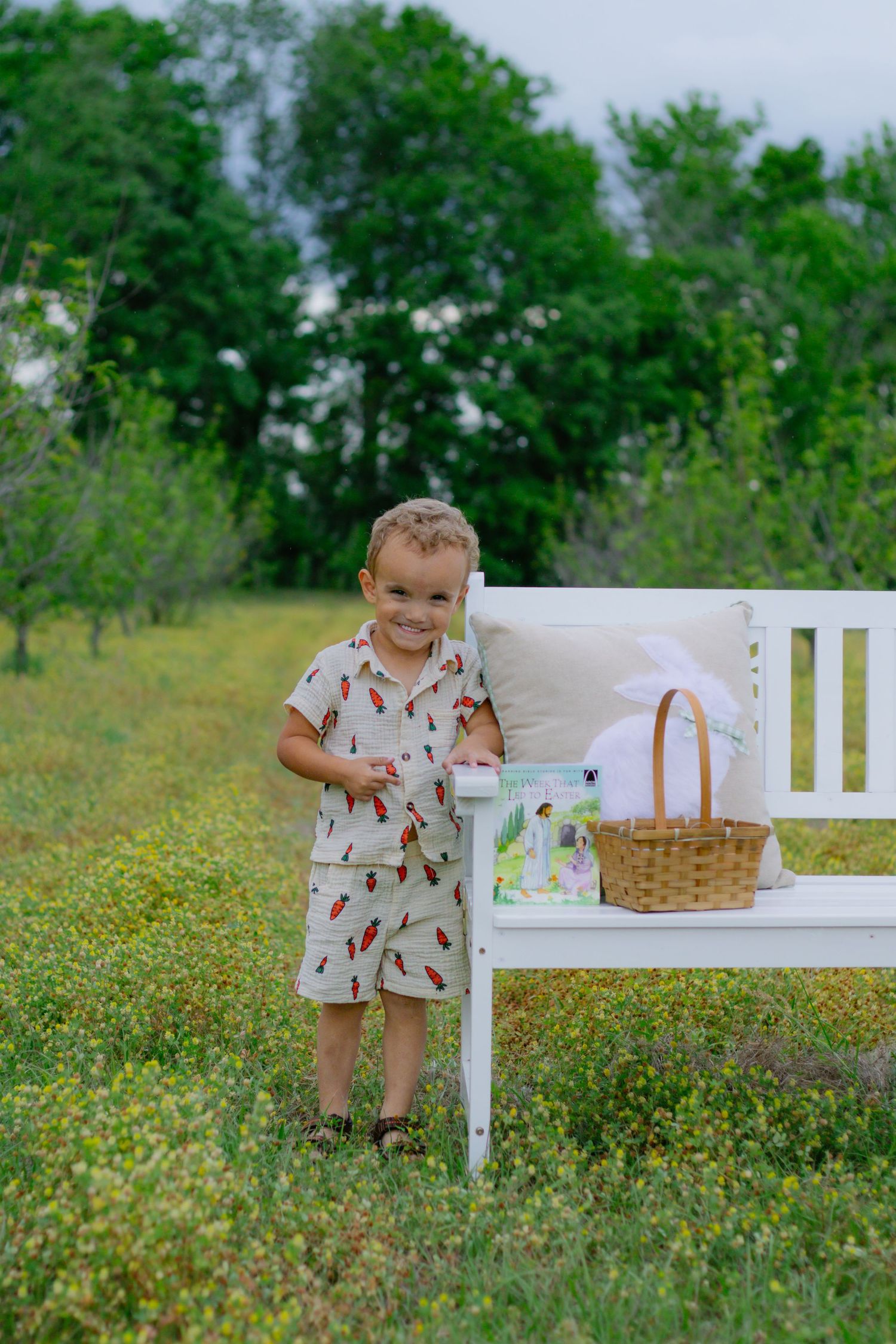 Happy young boy in carrot-print outfit stands smiling beside a white bench with Easter basket in a green meadow.