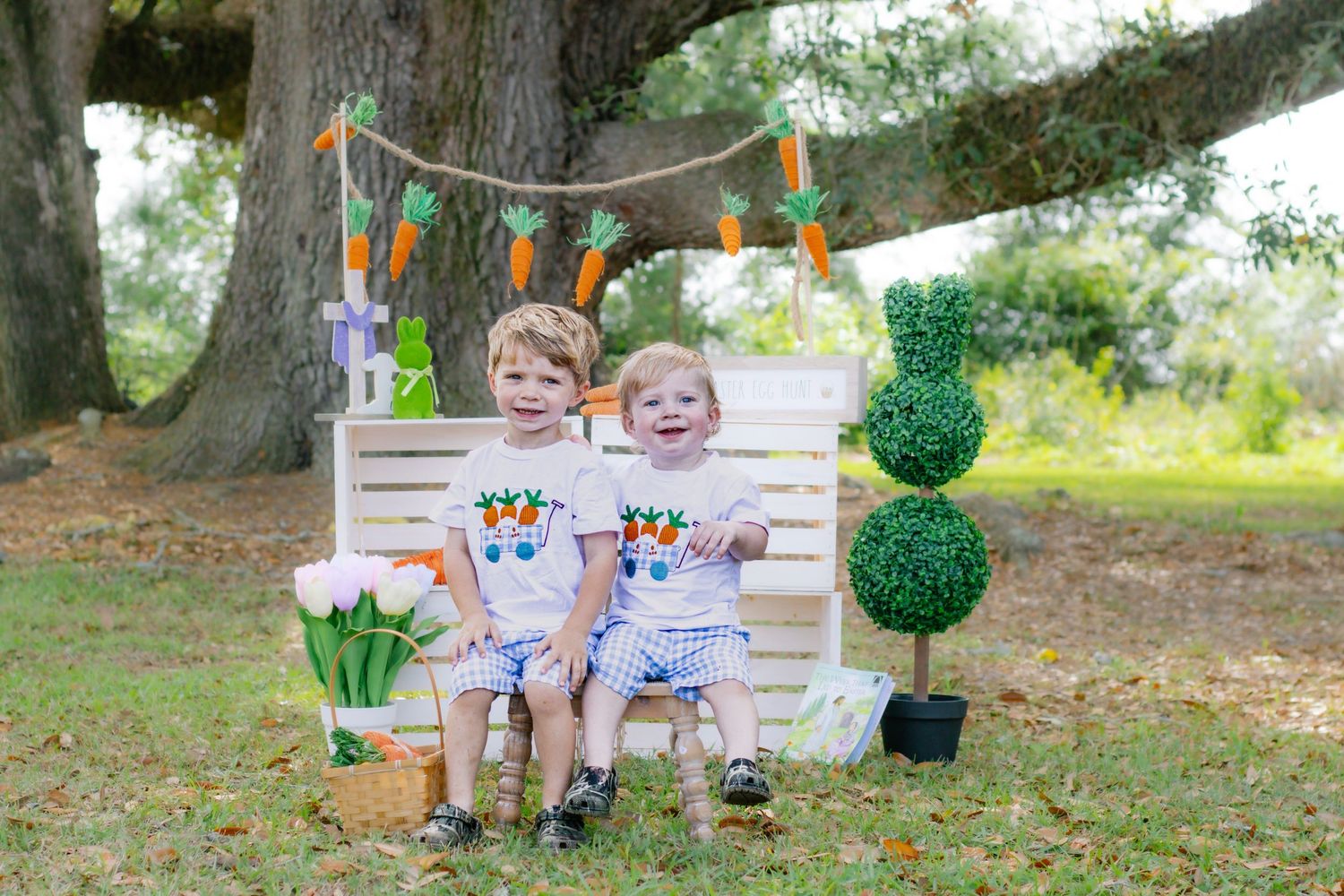 Two young boys in matching Easter outfits sit on white bench with carrot bunny decorations.