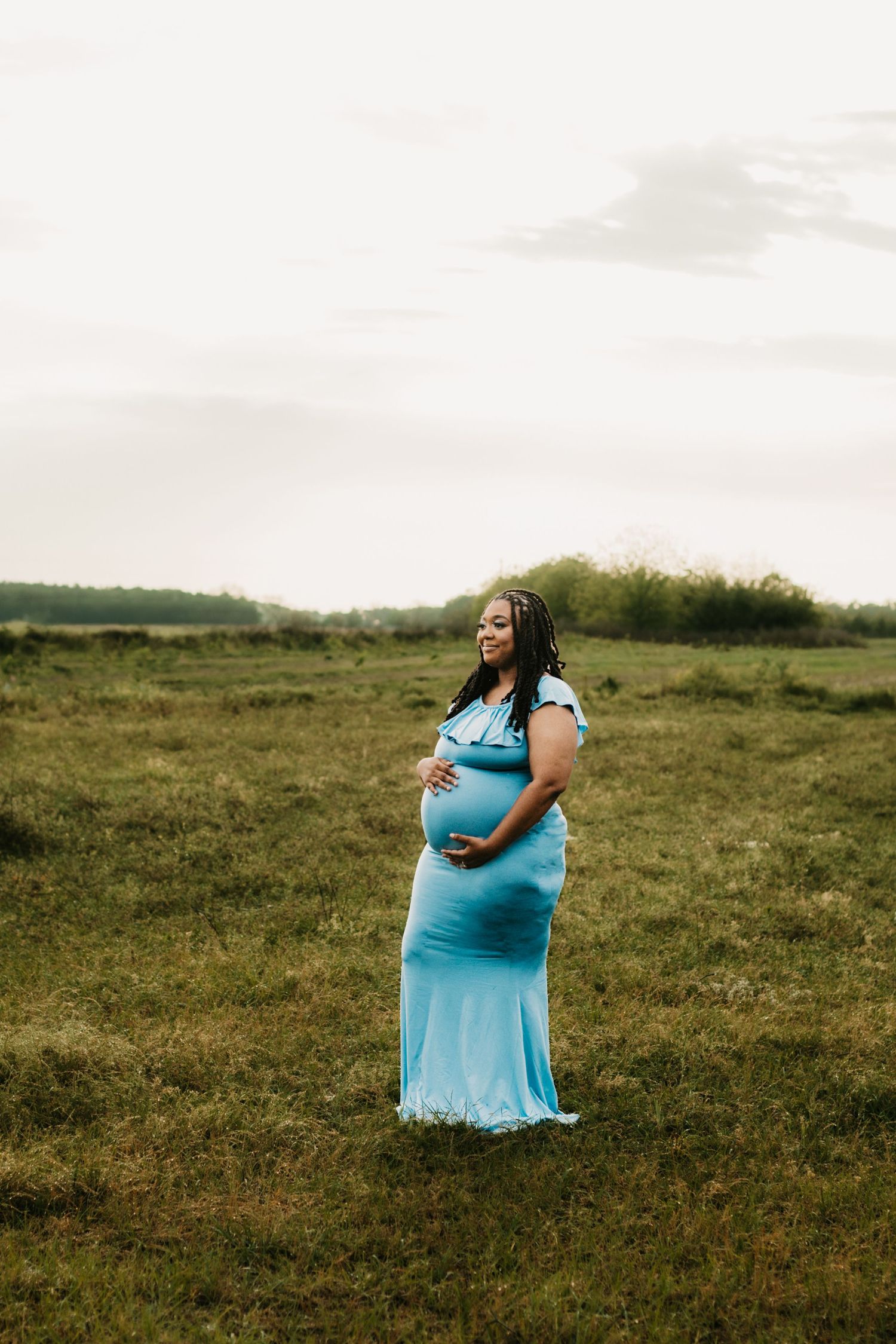 Glowing pregnant woman in light blue dress stands in green field cradling her baby bump.