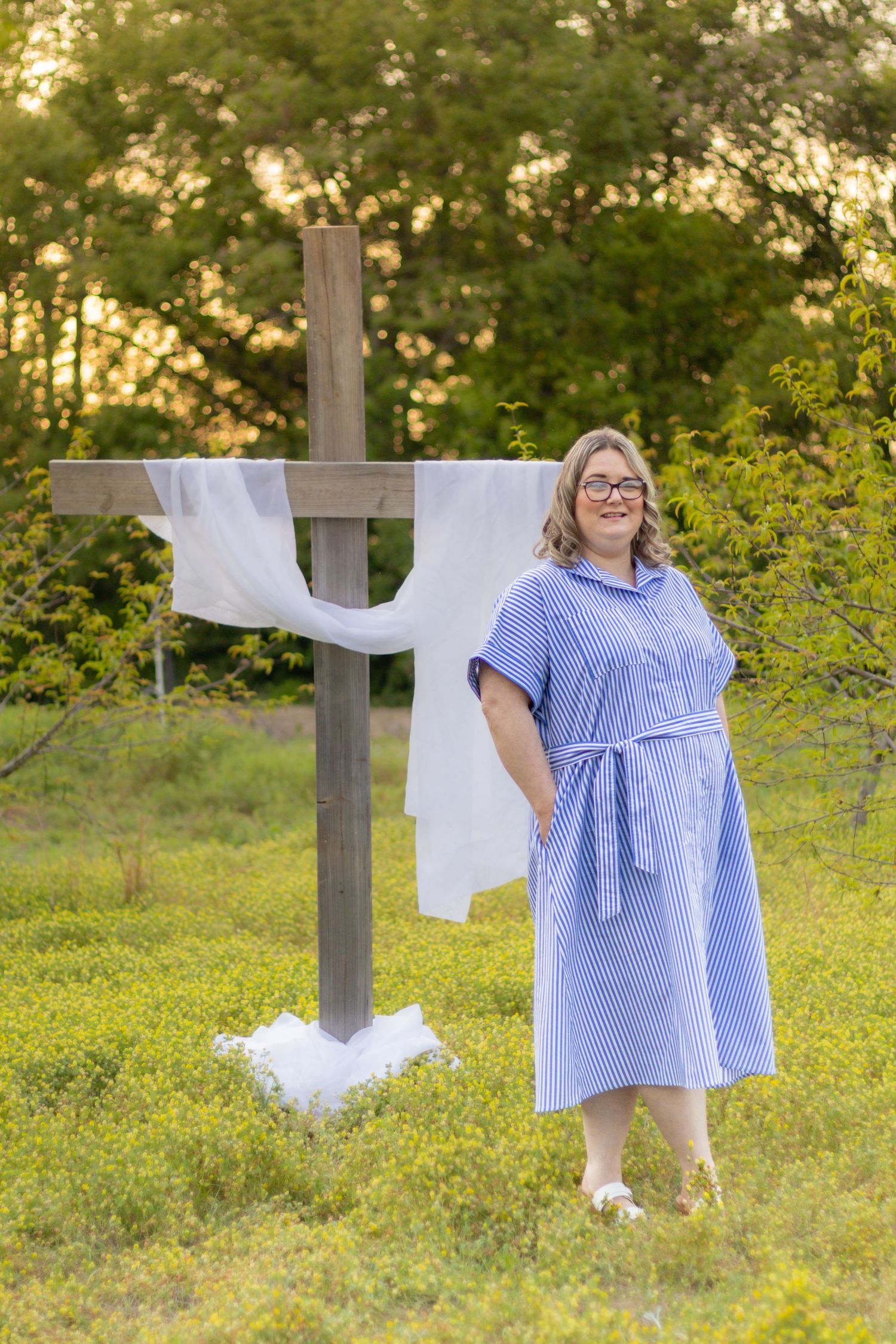 Smiling woman in blue striped dress standing beside a white-draped wooden cross in a green field at golden hour.
