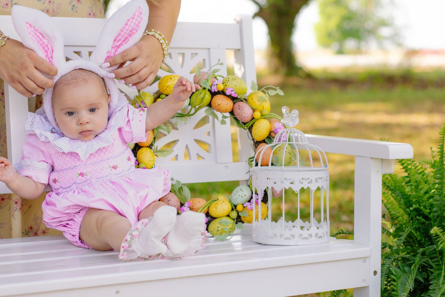 Baby in Easter bunny ears sits on white bench with colorful egg wreath and decorative birdcage outdoors.