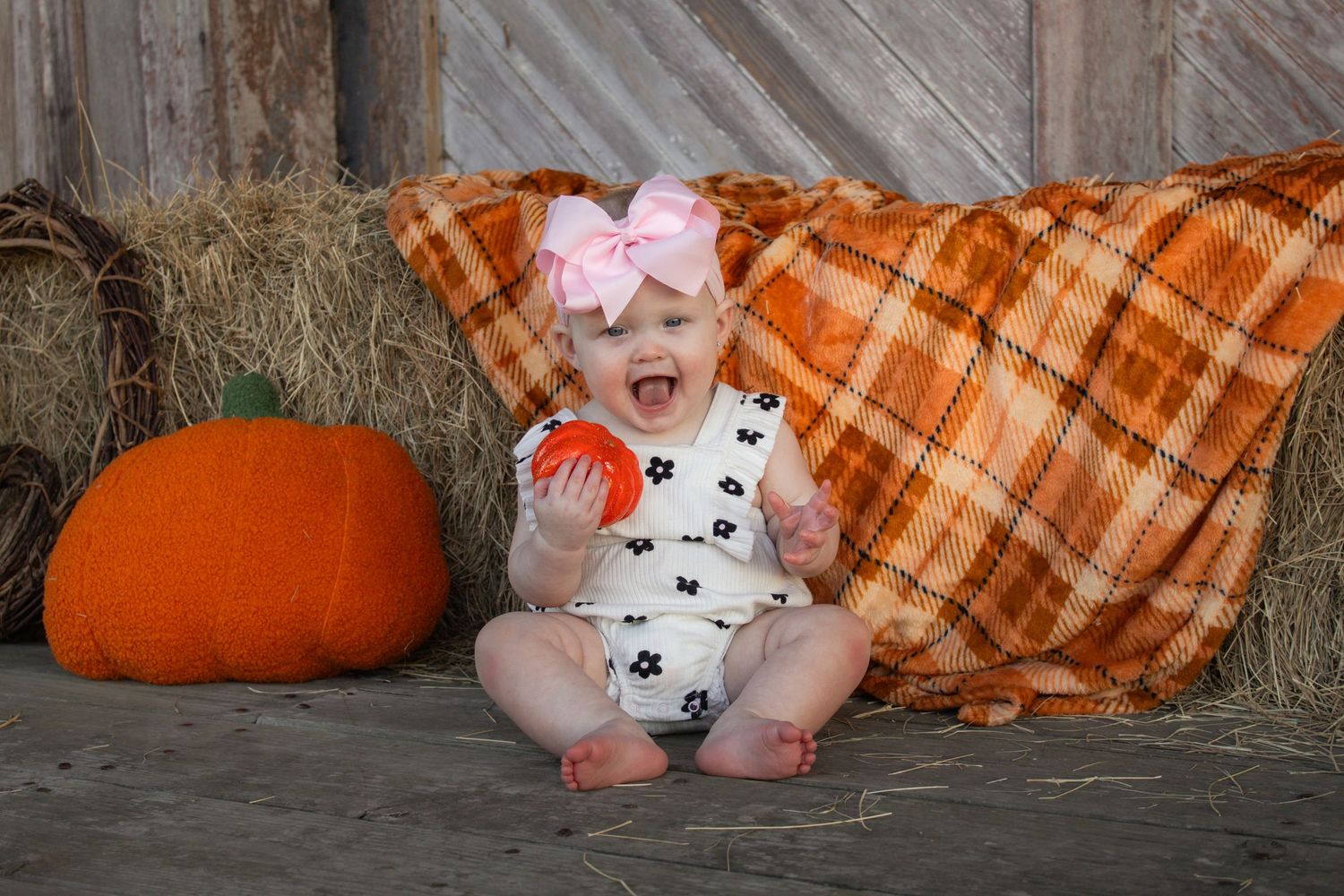 Smiling baby girl with pink bow sitting by hay bales and pumpkins in fall barn setting.