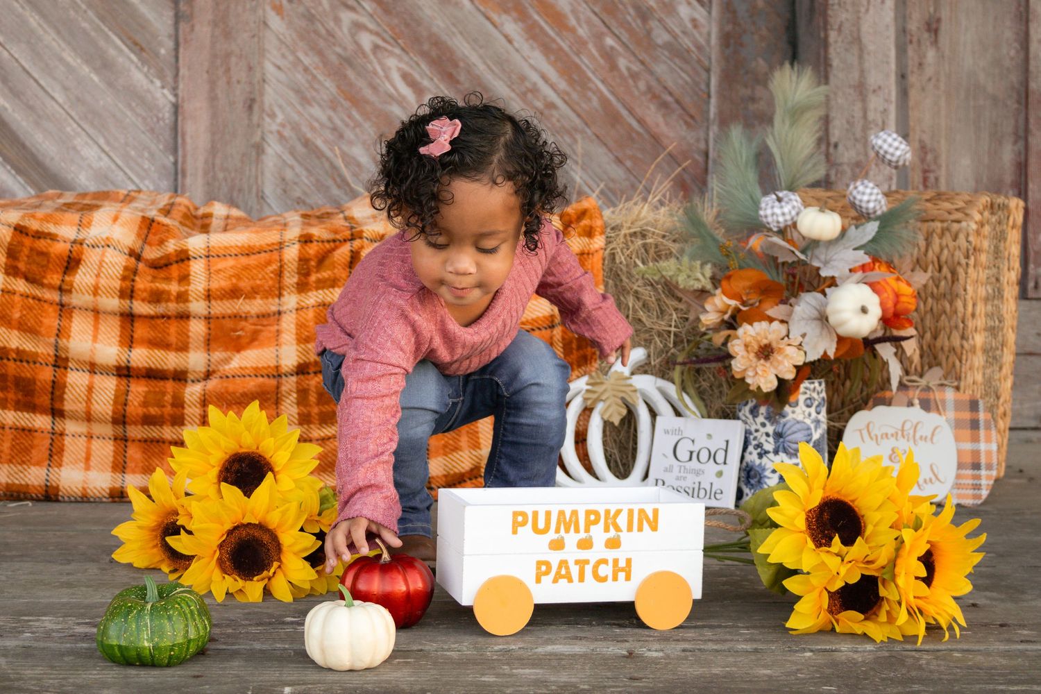 Toddler girl in pink sweater reaching for colorful pumpkins near Pumpkin Patch wagon with sunflowers.