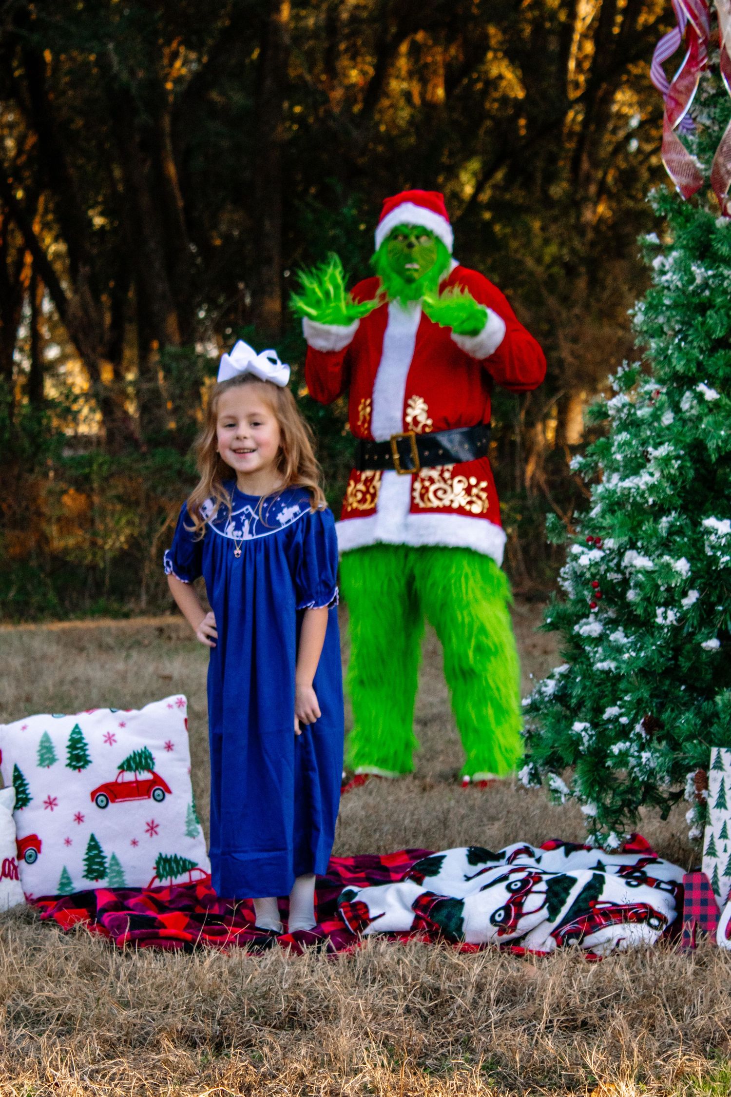 A smiling girl in a blue dress stands with the Grinch character waving beside a decorated Christmas tree.