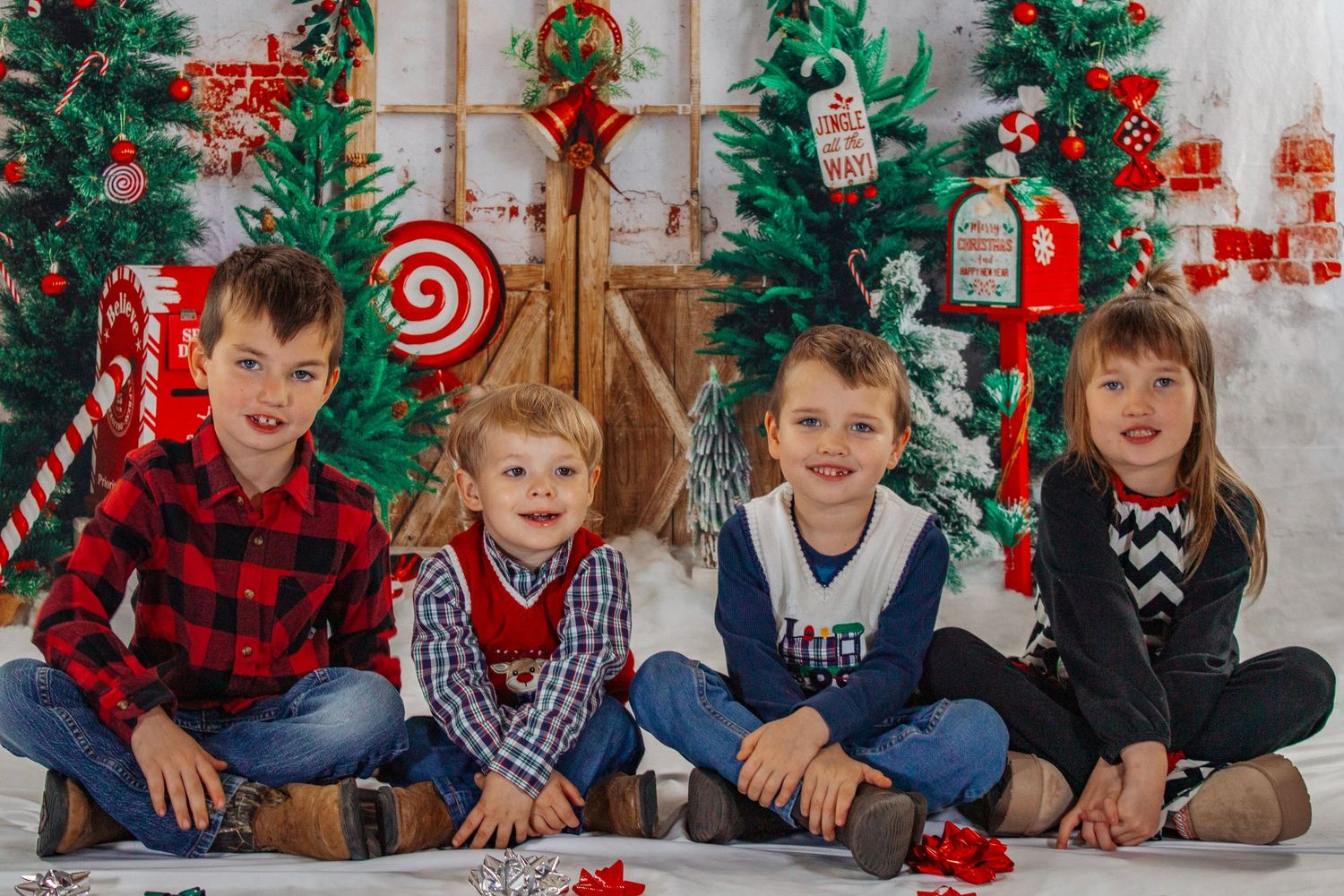 Four young children sit together smiling in front of a festive Christmas backdrop with colorful decorations.