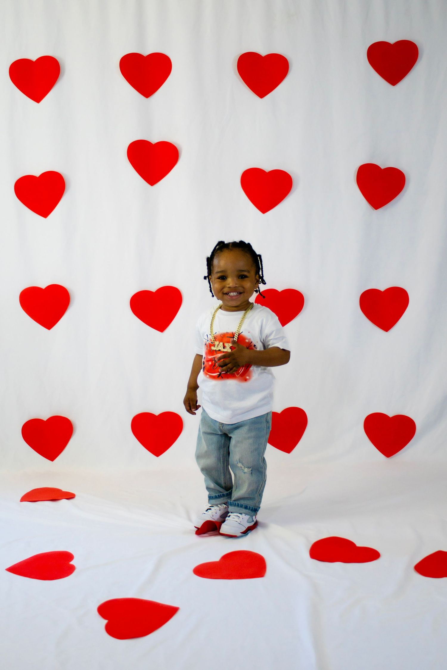 Toddler in white Valentine's Day shirt stands smiling in front of white backdrop with red heart decorations.