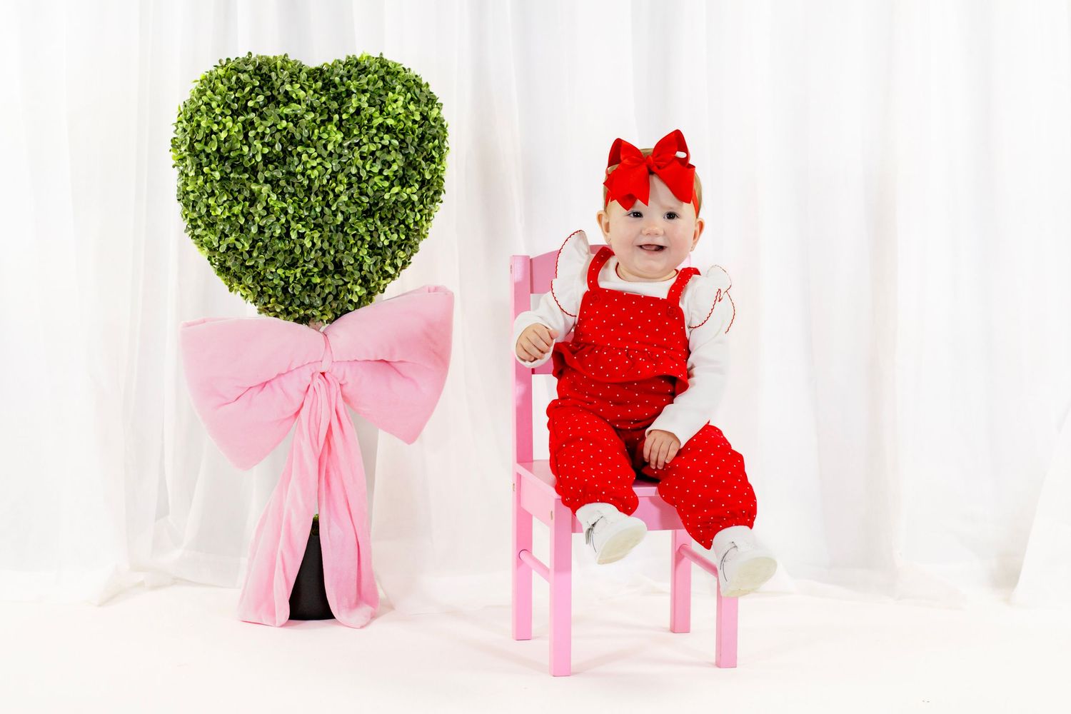 Baby girl in red polka dot outfit with red bow sits on a pink chair beside a heart topiary.