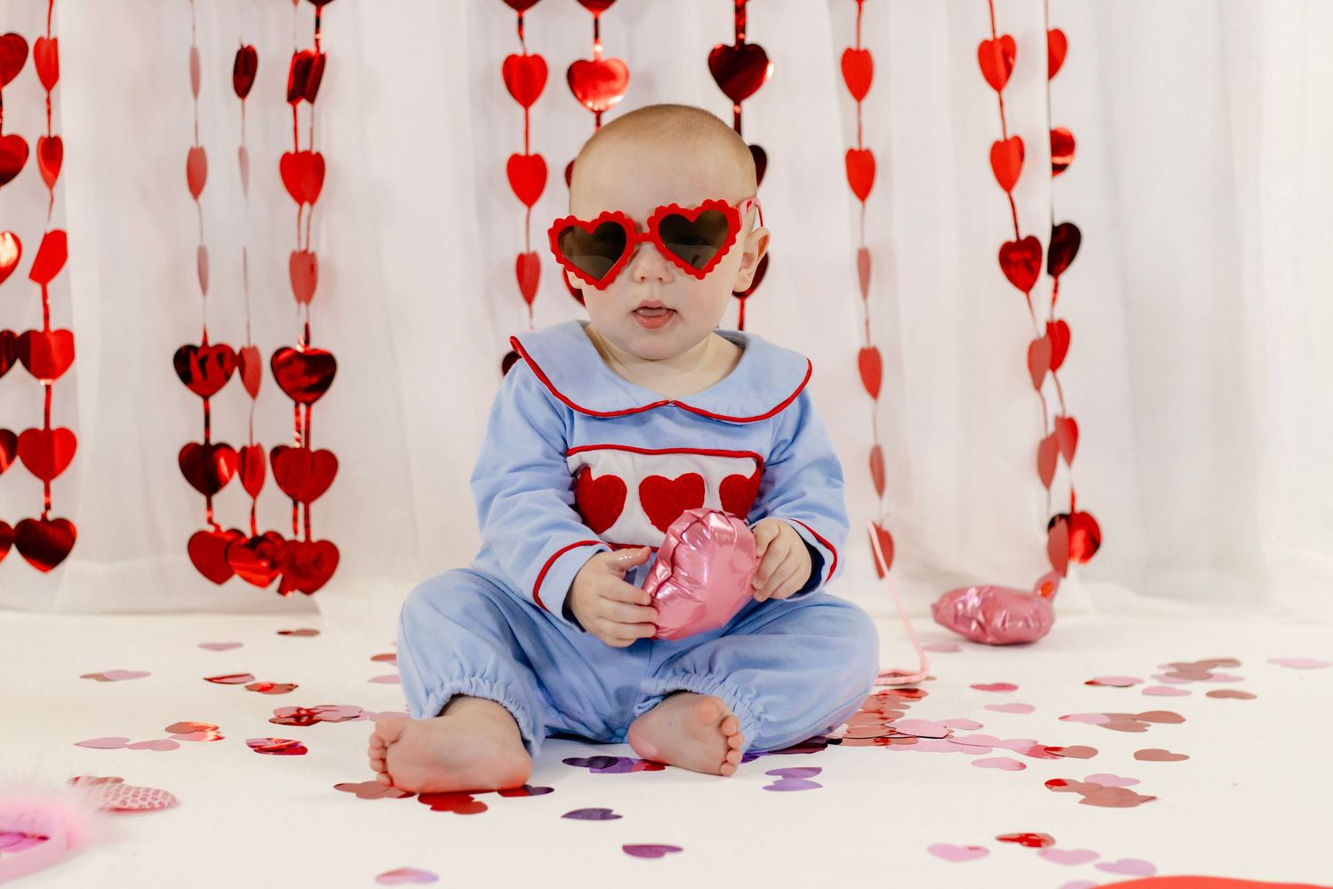 Adorable baby wearing heart-shaped sunglasses sits among Valentine's Day confetti in a blue smocked romper.