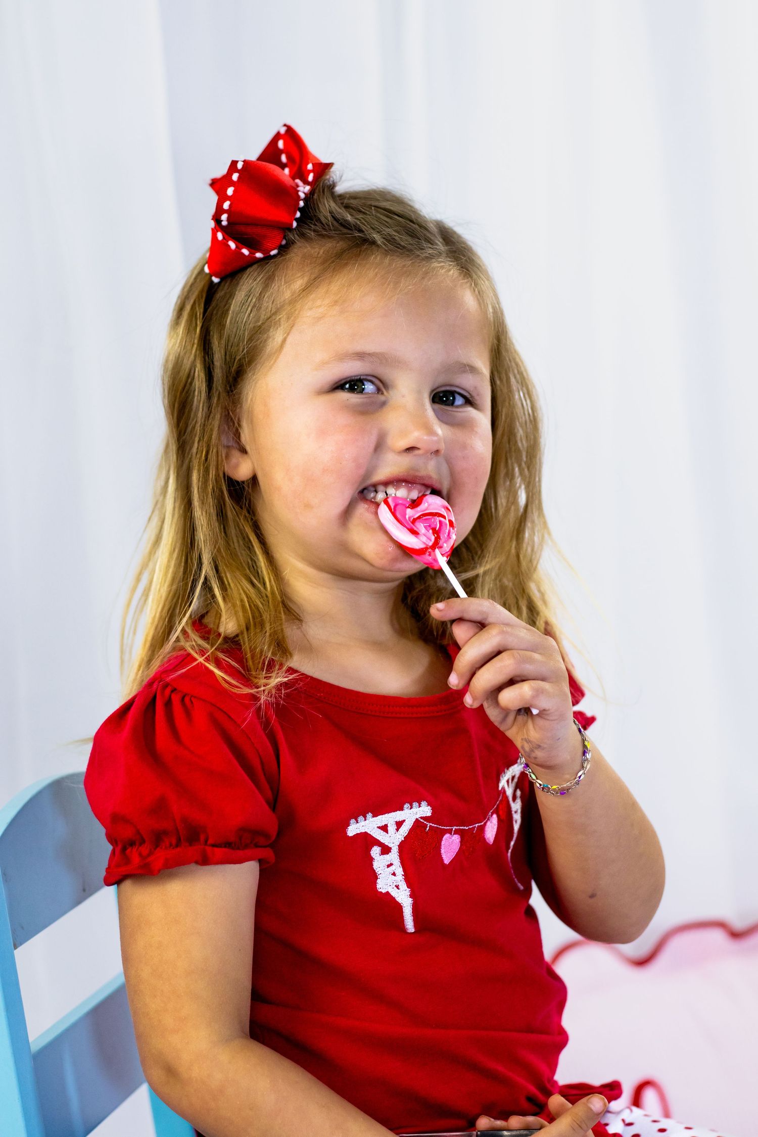 Young girl in red dress with bow eating pink heart lollipop in Valentine's Day photo session.