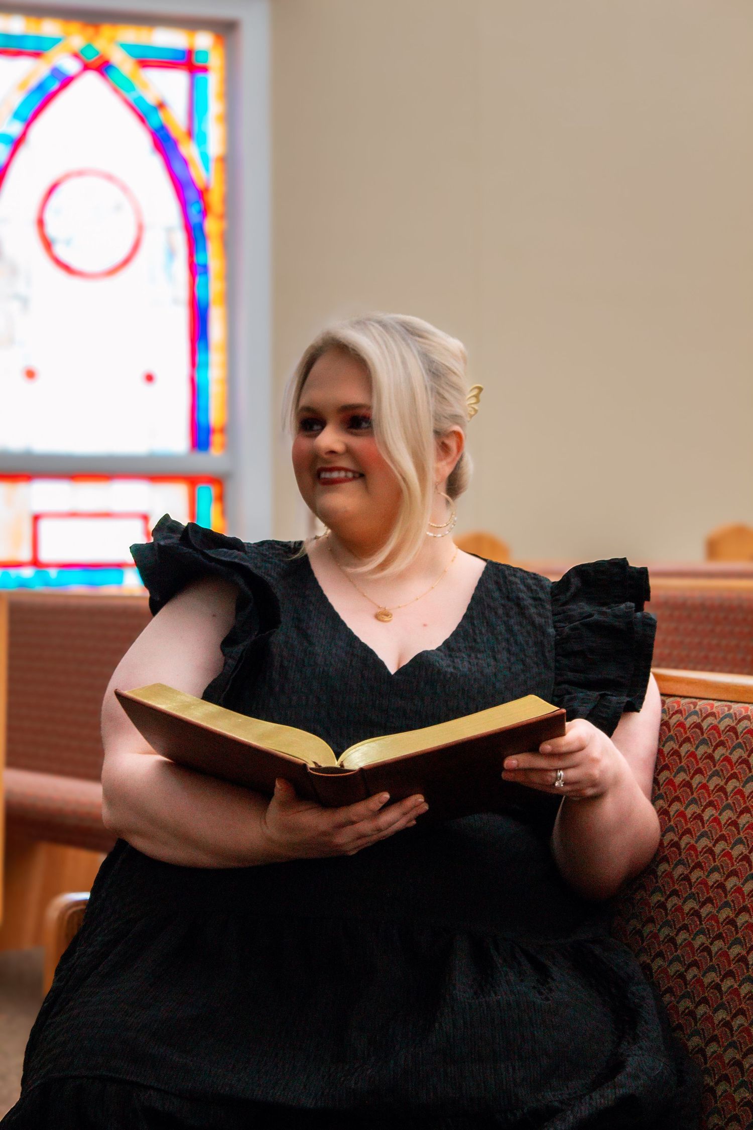 Smiling blonde woman in black ruffled dress reading a Bible in a church pew with stained glass window.